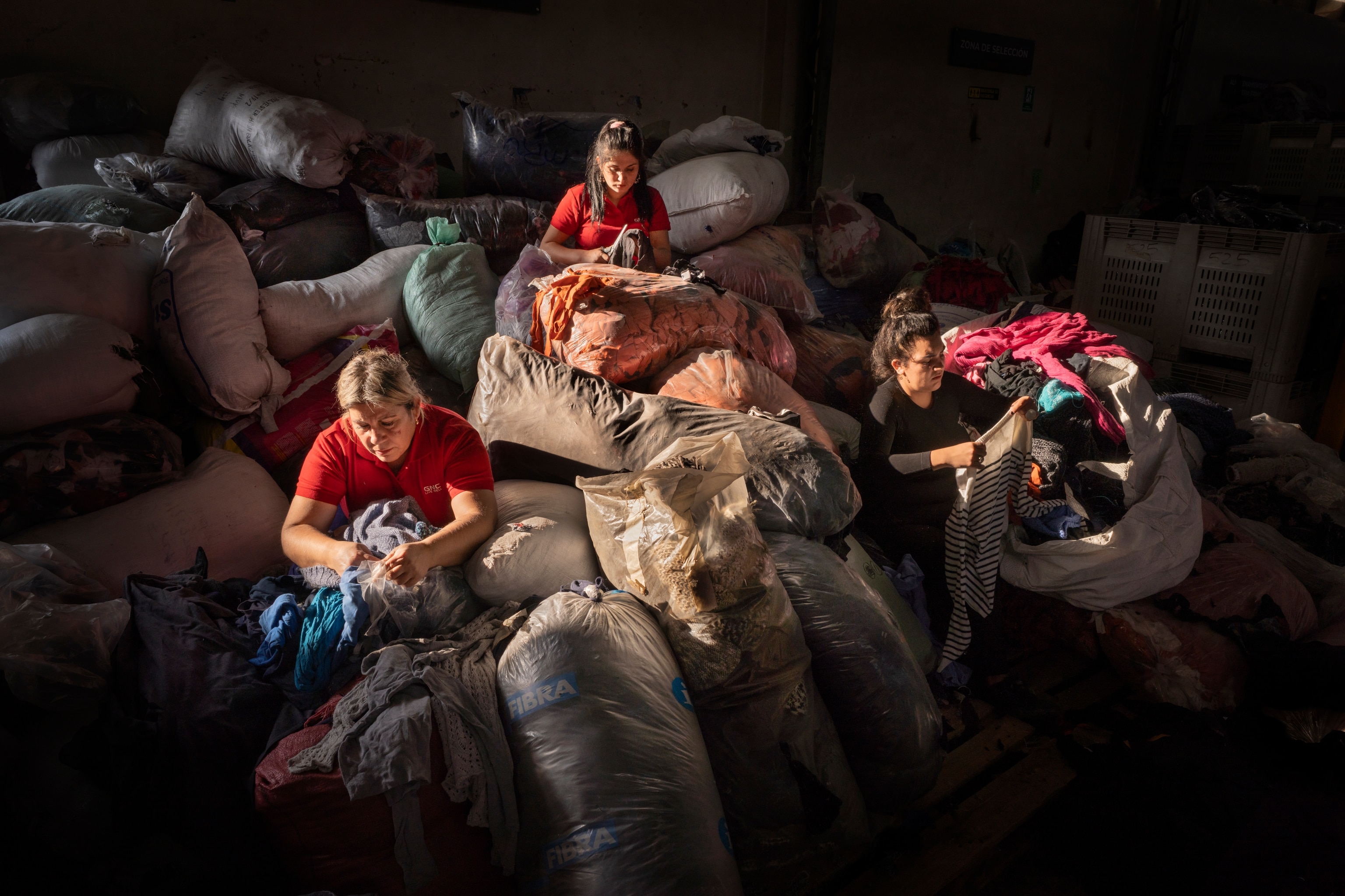 three women are seated with dozens of trashbags of clothes surrounding them. They are each going through one bag each.