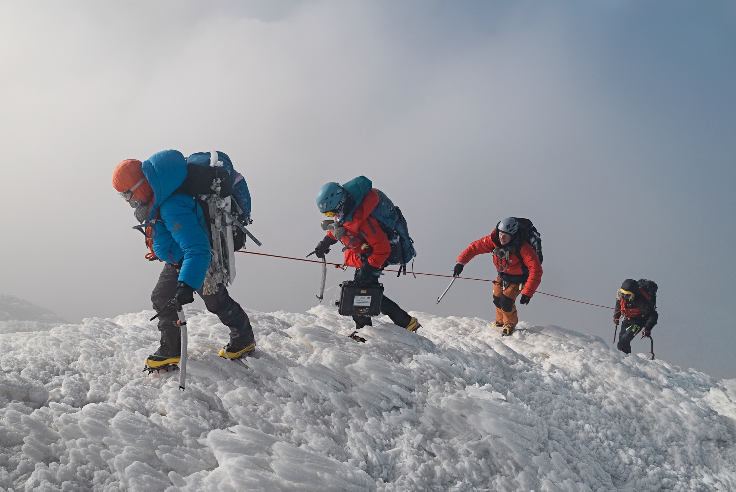 Four people connected by the rope, waring crampons and bright jackets, carrying large backpacks as working uphill.