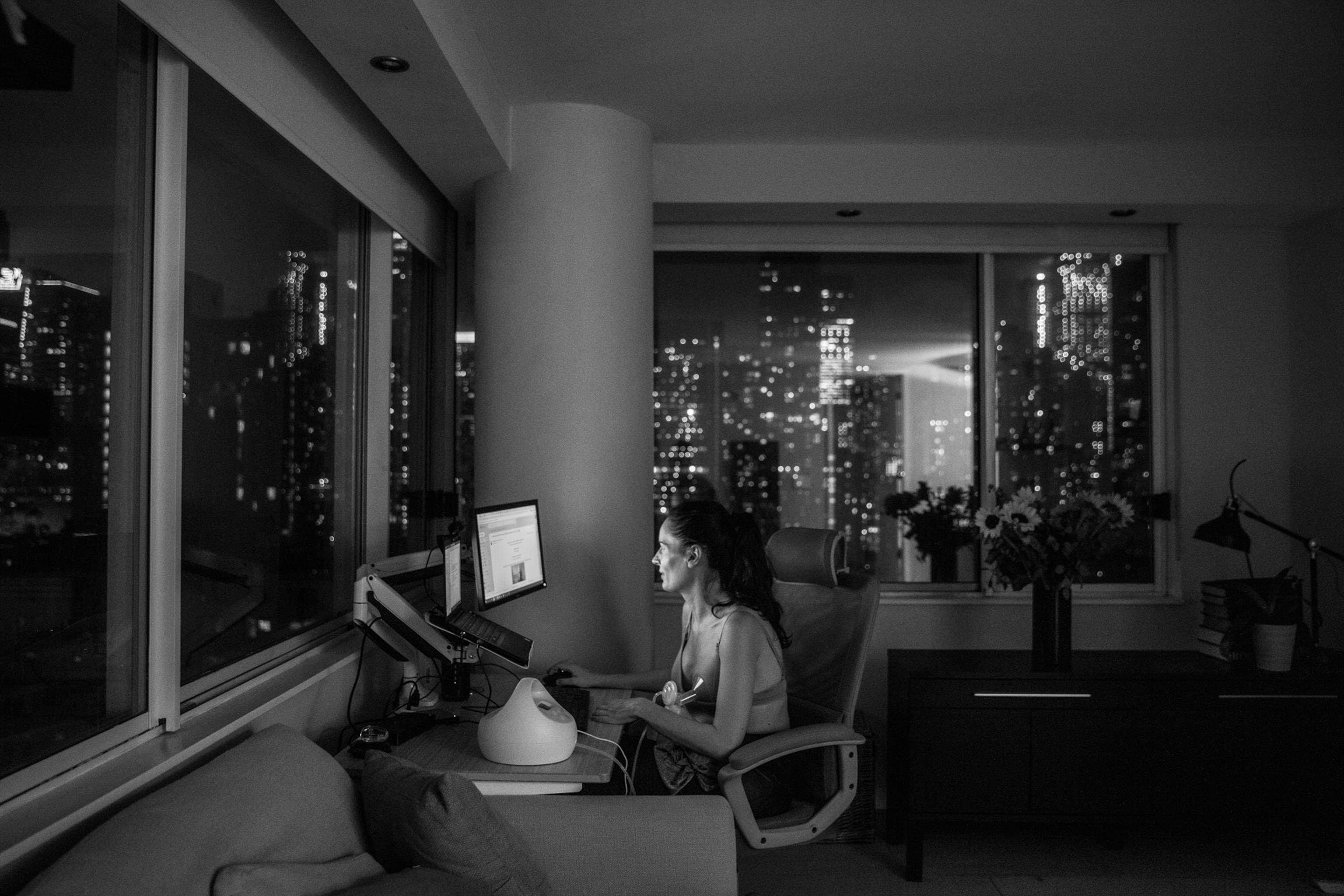 Woman working on computer in corner room with open view of big city at night.