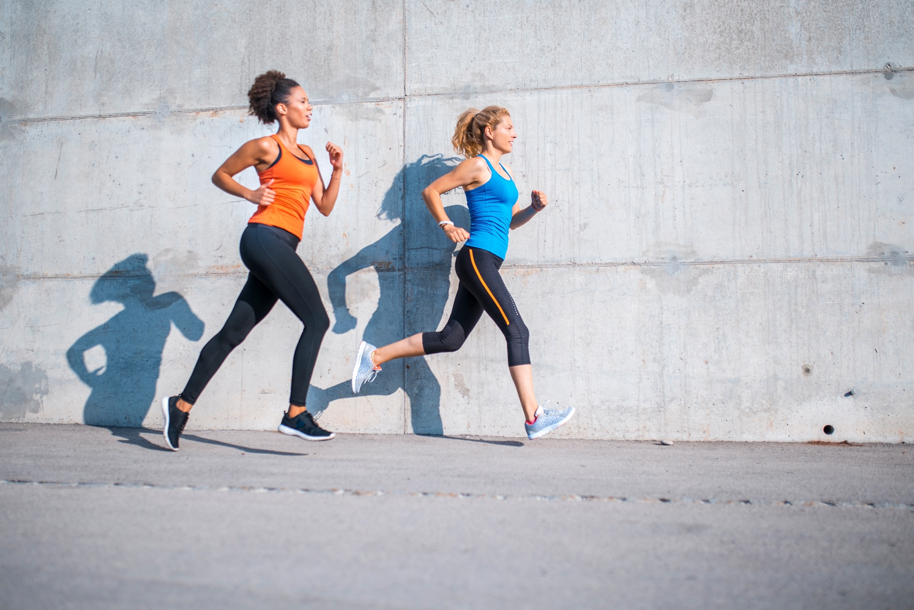Two wmen the one closer to the foreground had a darker tone and curly hair pulled back out of her face with an orange tank top, the women in the back has paler skin with a loose pony tail a blue tank top with light blue sneakers.