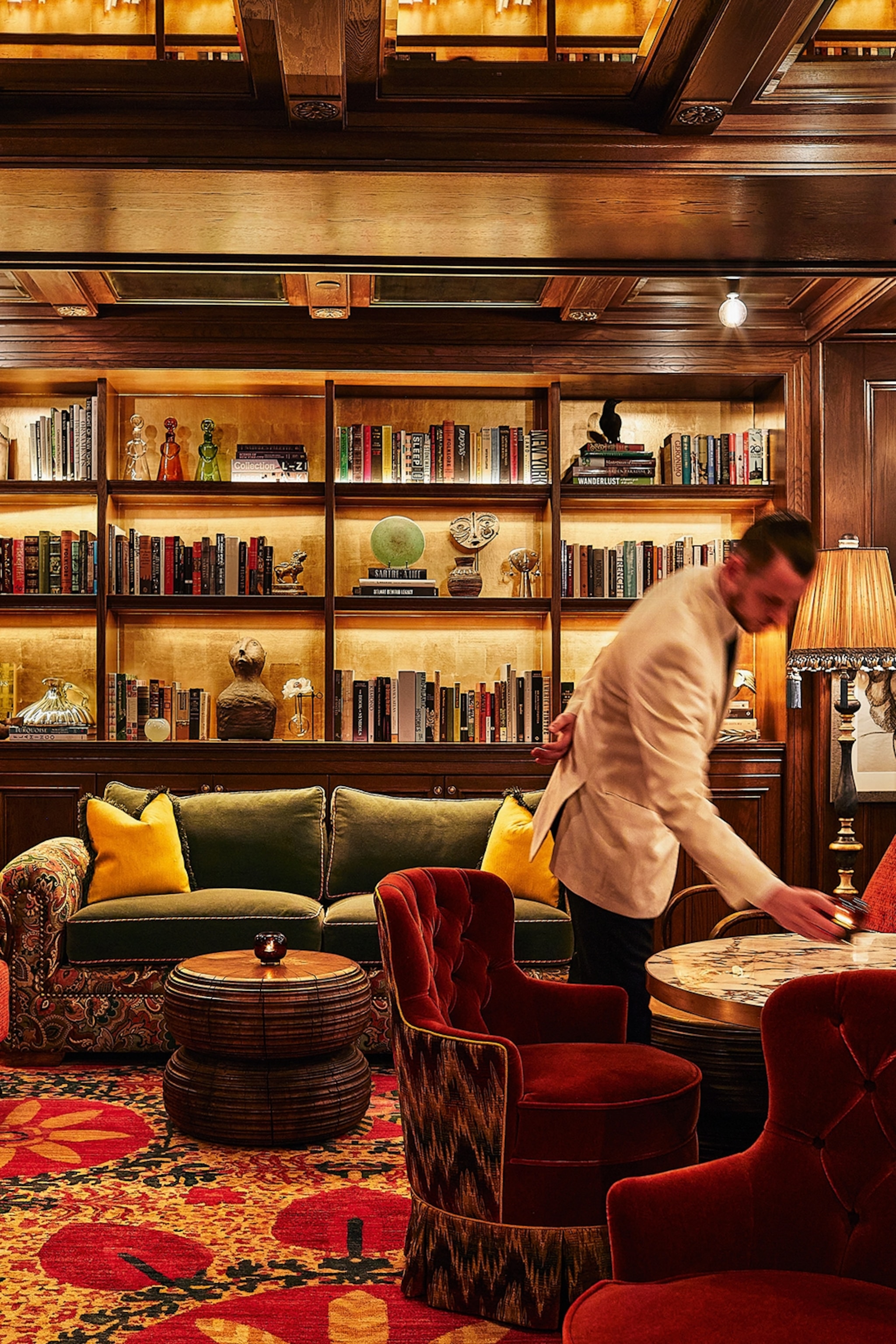 Waiter clearing a table in a glamorous, elaborate library surrounded by velvet furniture and books
