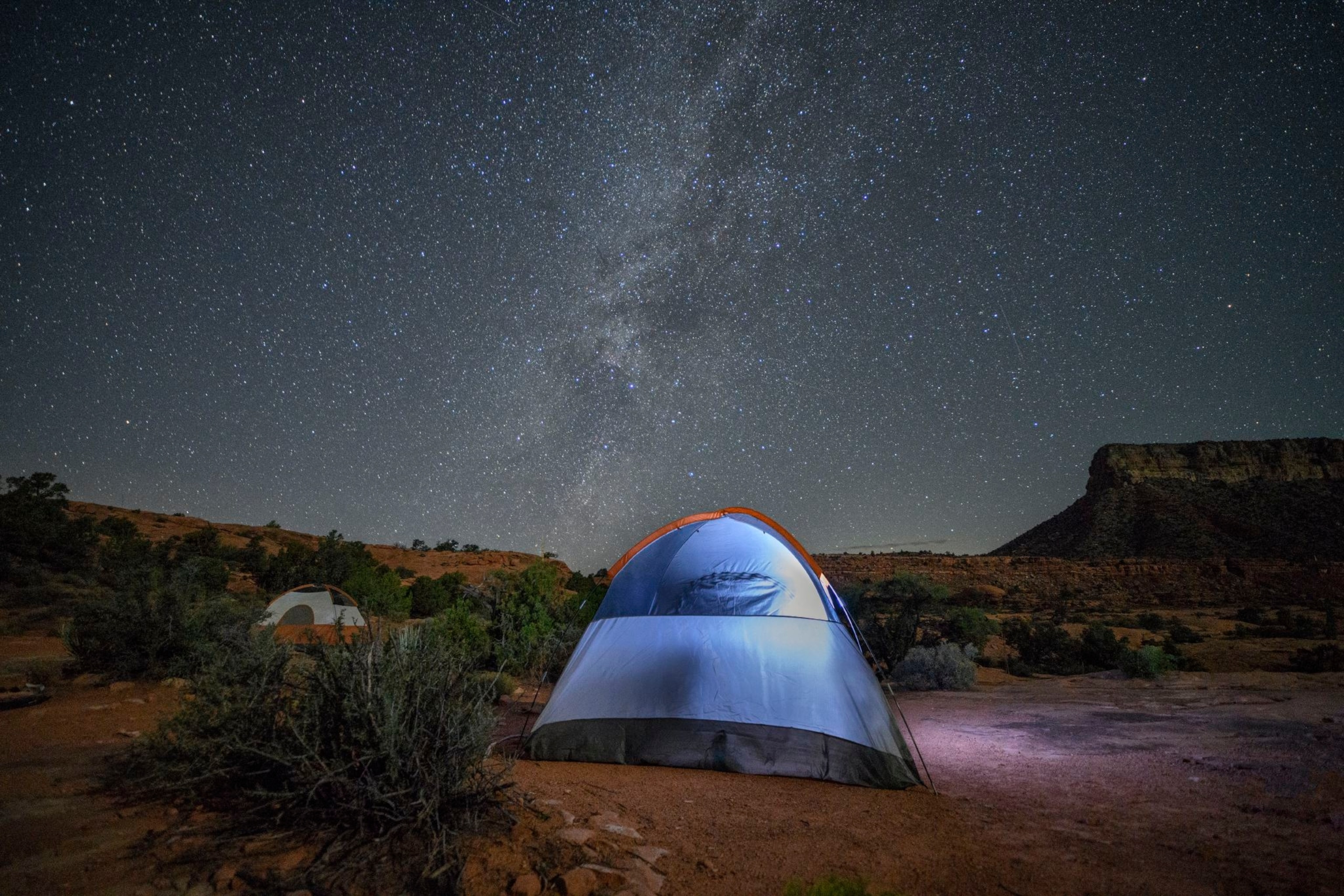 Two tents sit among desert brush, illuminated from within as they sit under the Milky Way hanging in the night sky.