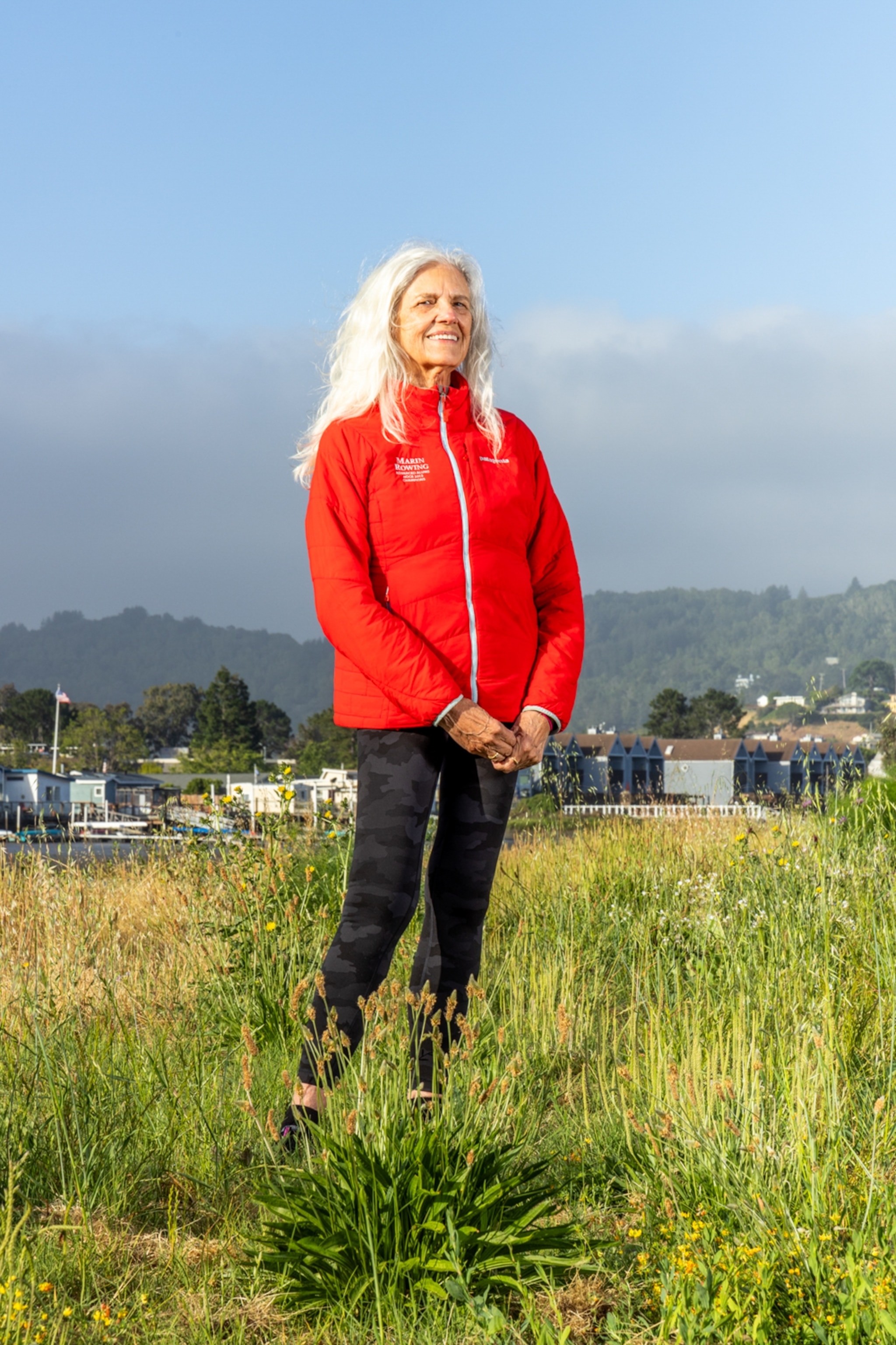 A woman with long white hair with red jacket in a green field and a soft blue sky.