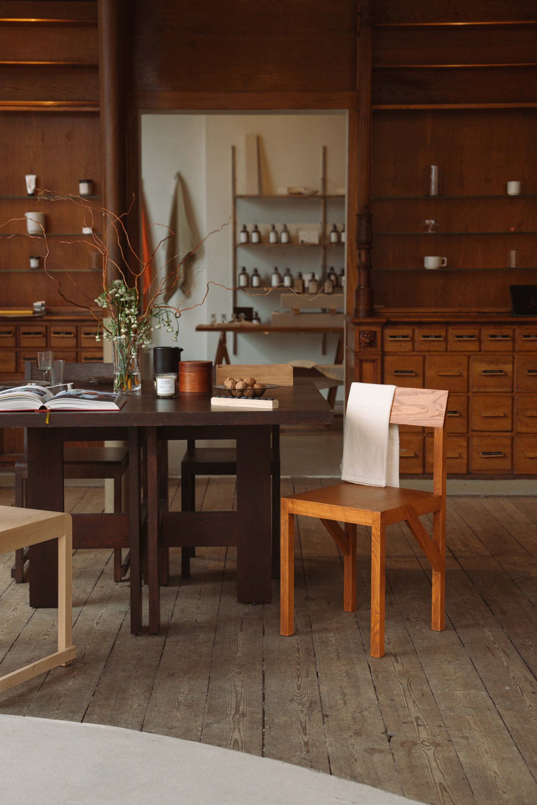 A modern table and chairs sit in the foreground. On the wall behind them are shelves and drawers. A walkway through that interior separating wall shows another room in the background.