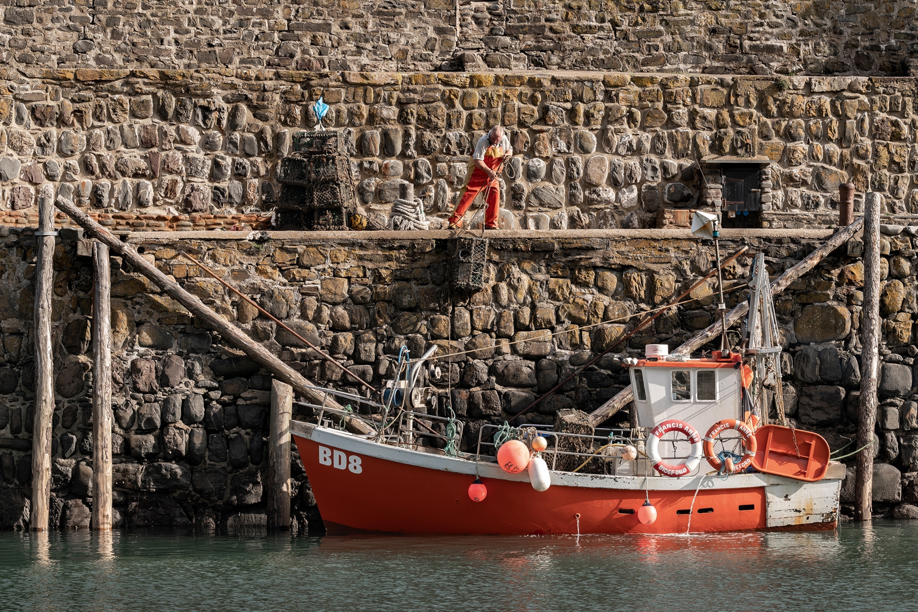 A fishing boat in Devon, England is docked against a rocky wall, with a fisherman lowering in crates from above.