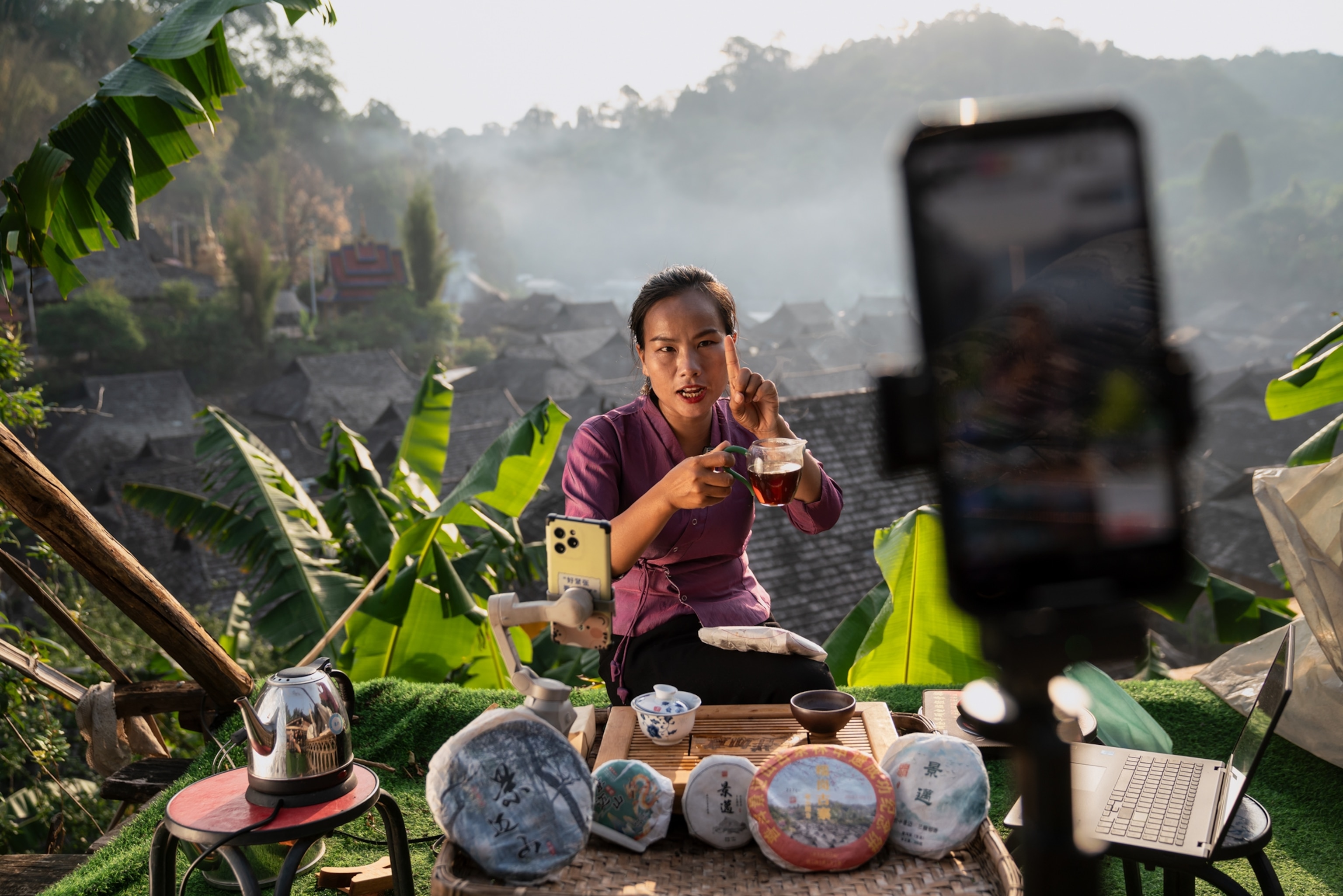 A woman sits holding a cup of tea in front of a table of packaged tea cakes. A phone records her in the foreground.