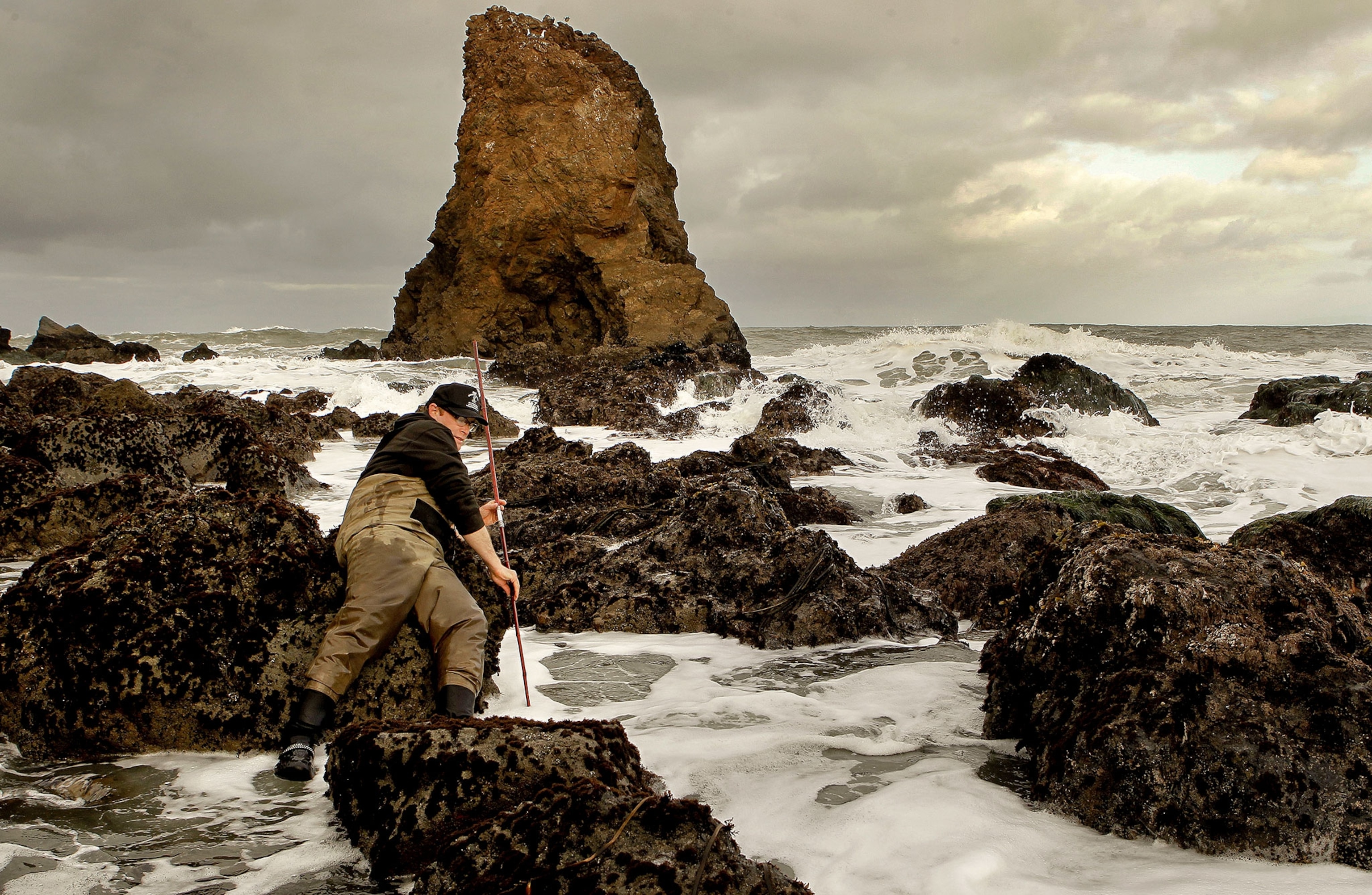 A man with a pole stands with boots and waders in a tidal pool with waves and rocks