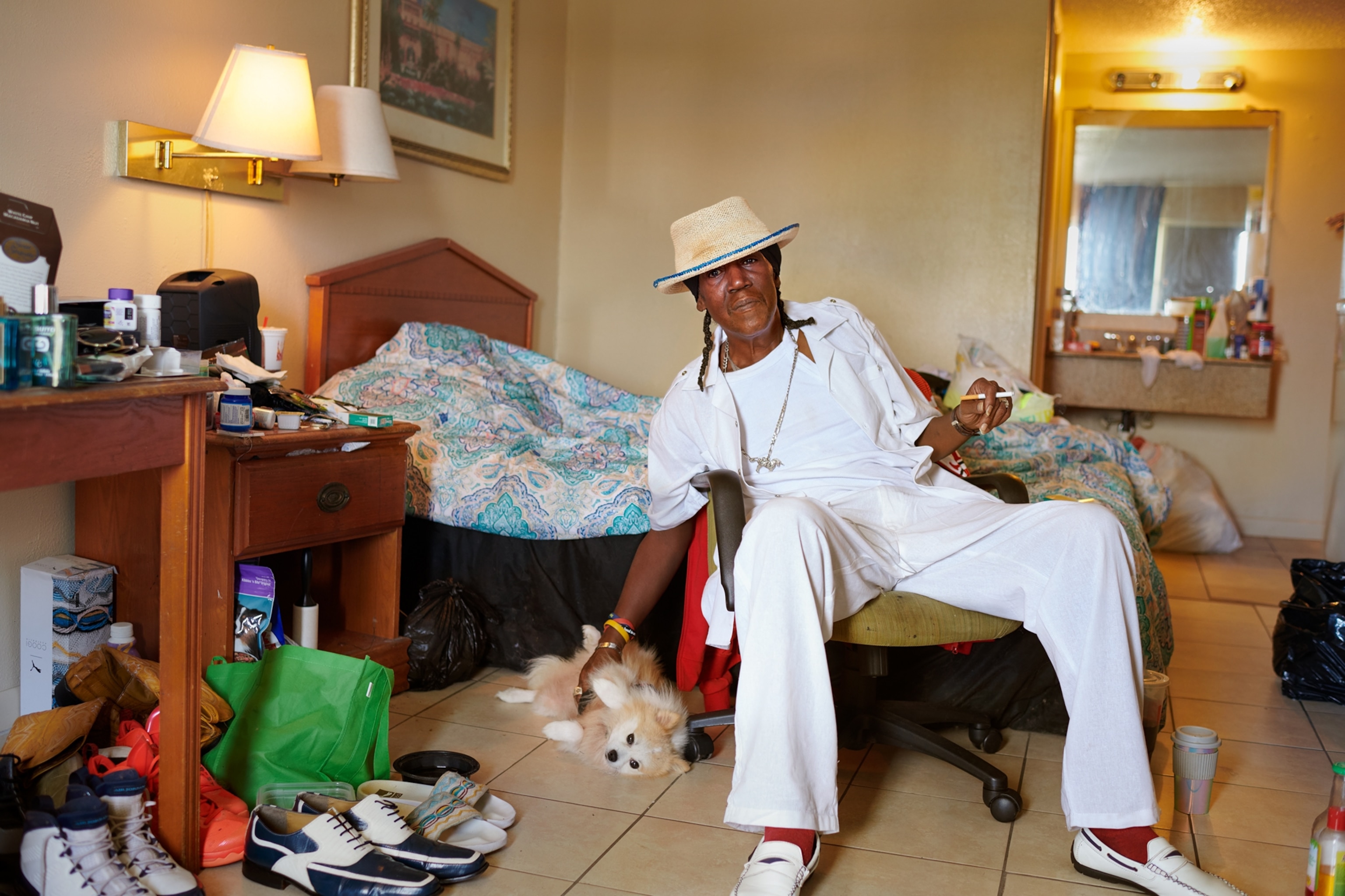a man dressed in all white and in straw hat sitting in chair and patting a small dog.