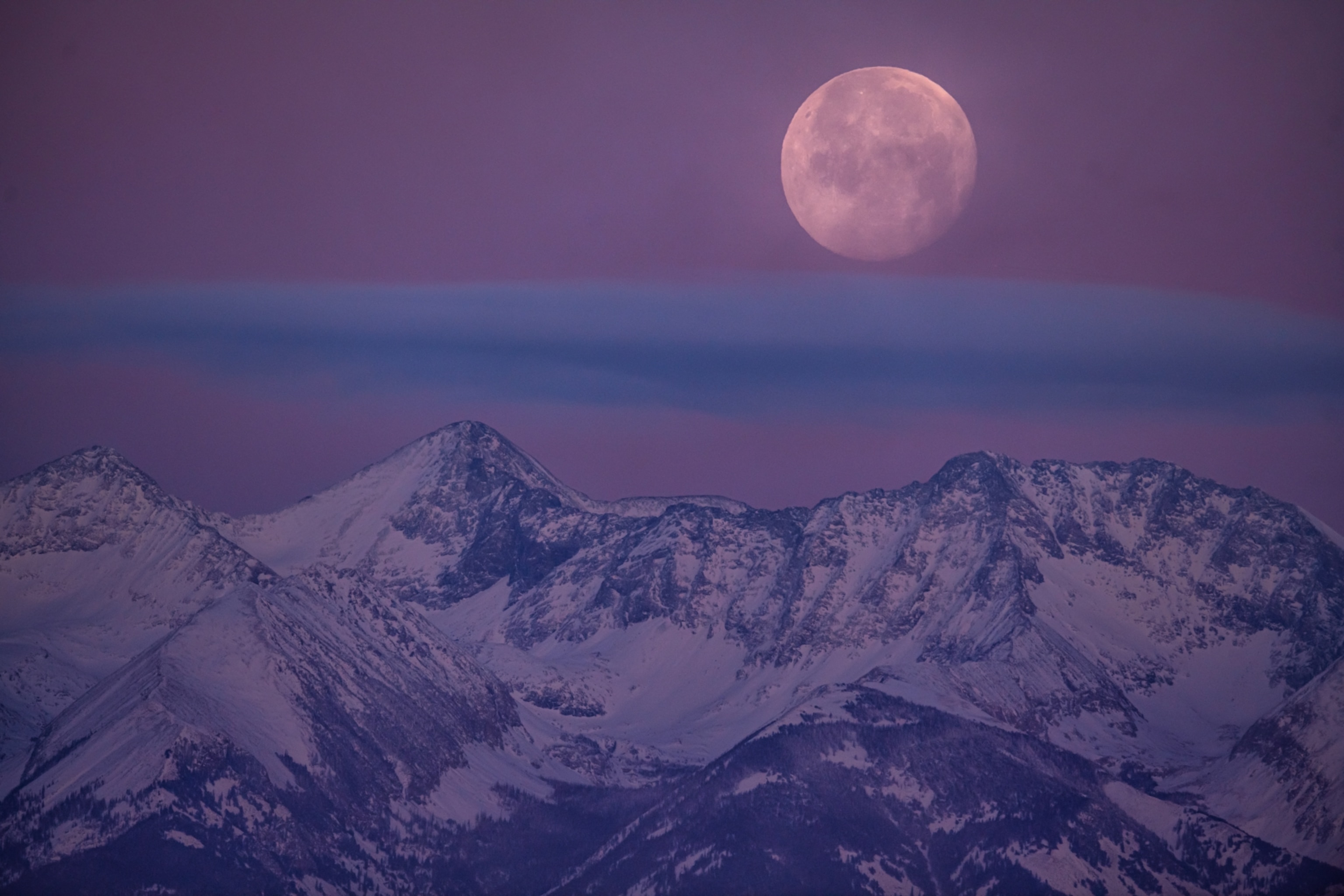 A full moon seems to rest atop a saucer-like cloud hanging in the purple sky beyond the snow-covered Sangre De Cristo mountains in Colorado.