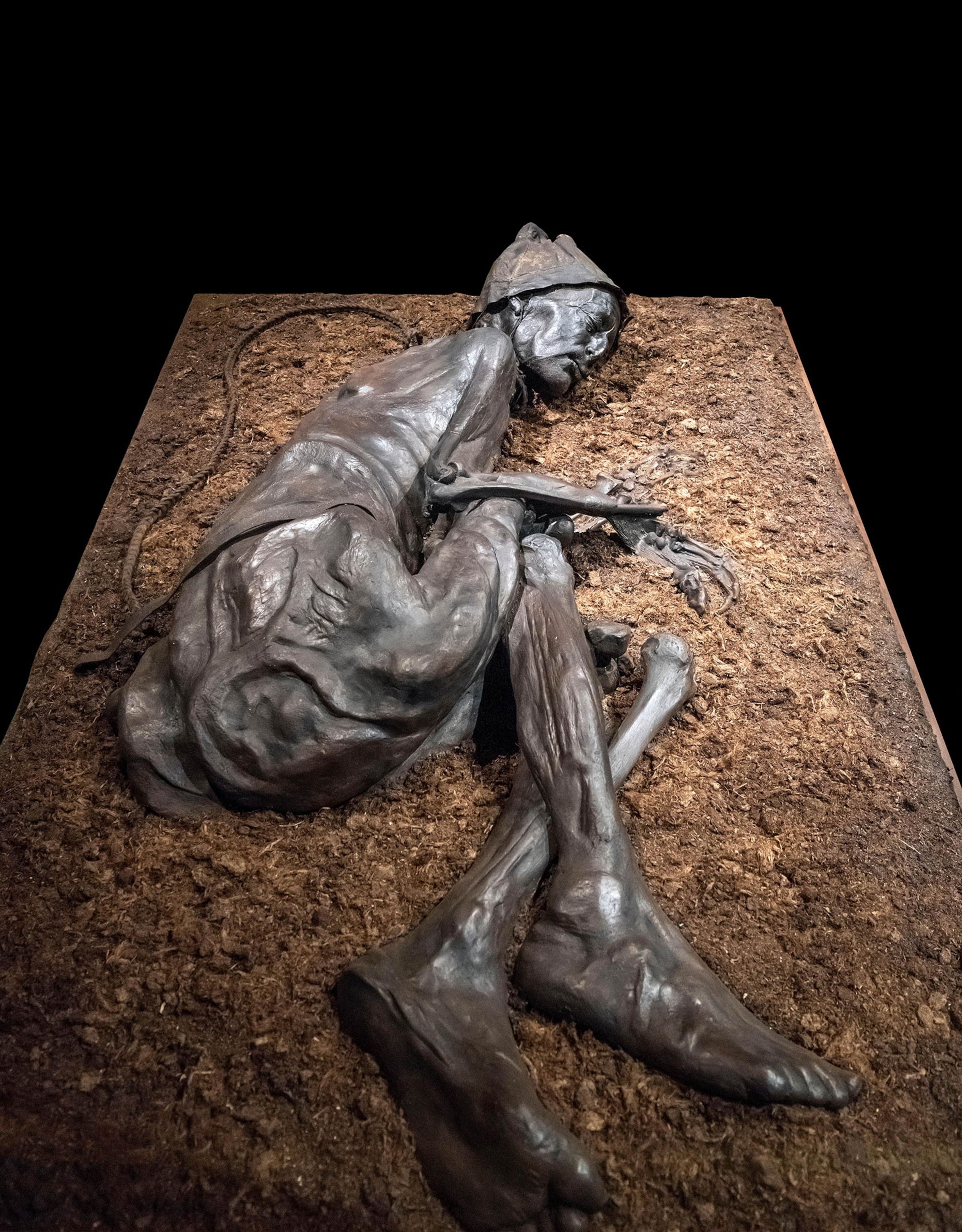 A brown, bronze like mummified body of a man laying on his side in the fetal position on a square pile of brown earth and soil, with a braided rope around his neck against a black background.