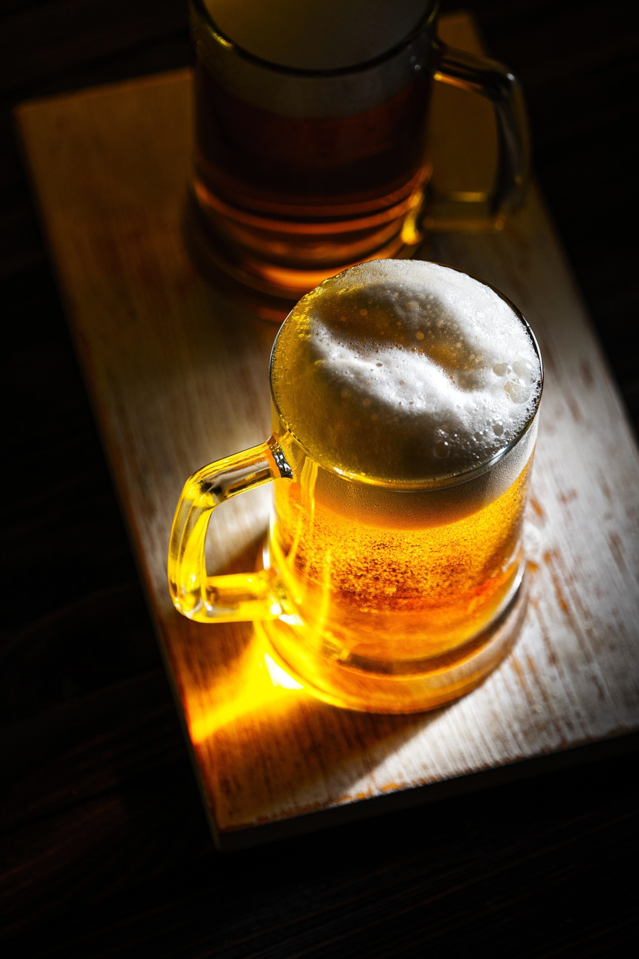 Two glass mugs of light beer with white foam seen from above, resting atop a dark wooden table.