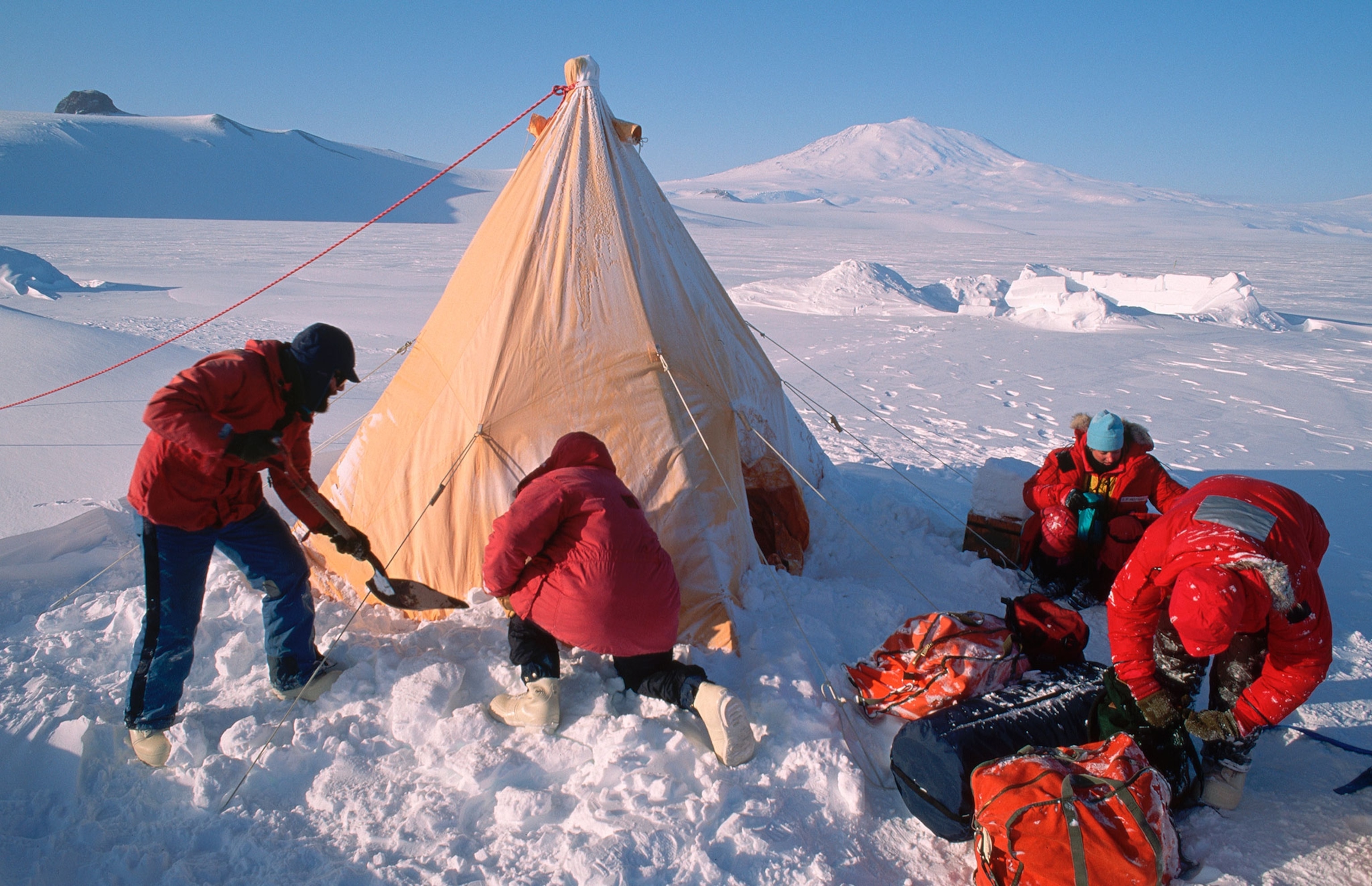 A group of men, dressed in red, winter coats, snow pants, and boots, pitch a tent in a snowfield during compulsory survival training near the US Base at McMurdo, Antarctica.