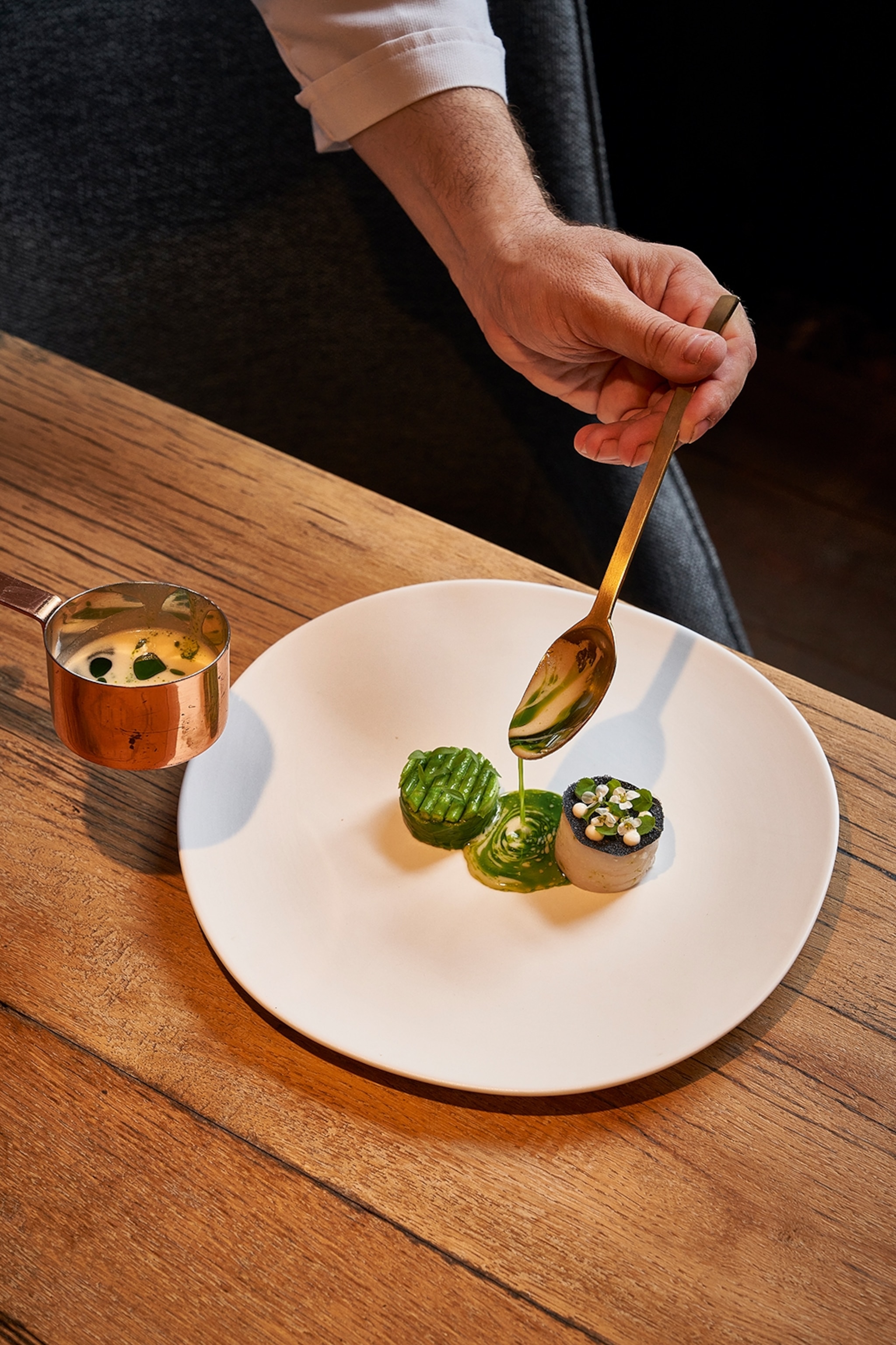 A large flat plate with two small medallions of food in the center, a marbled creamy sauce with herb oil is being poured in between.