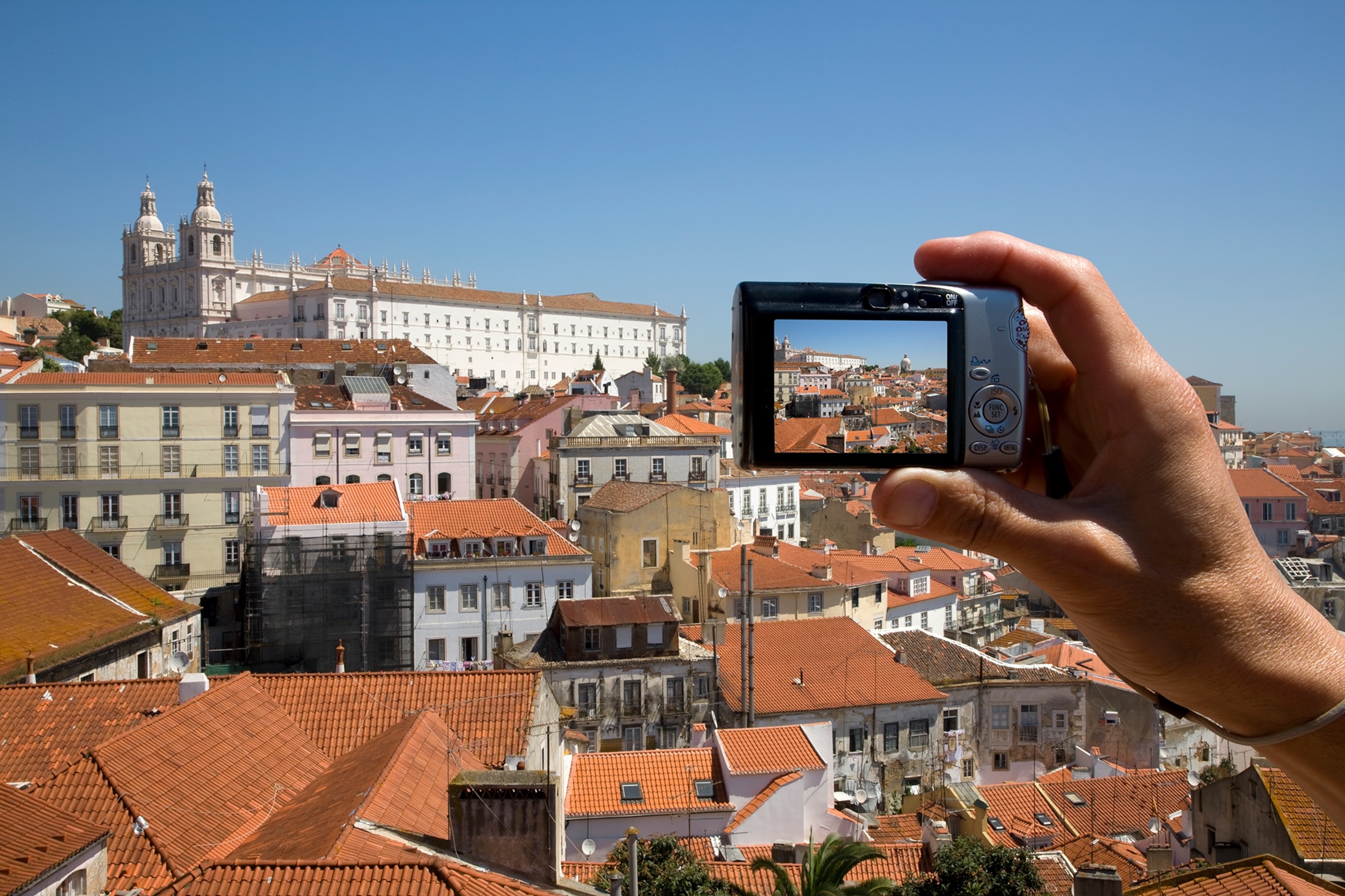 A mans hand holding a small silver camera with a strap photographing a city with terracotta colored roofs under a blue sky.
