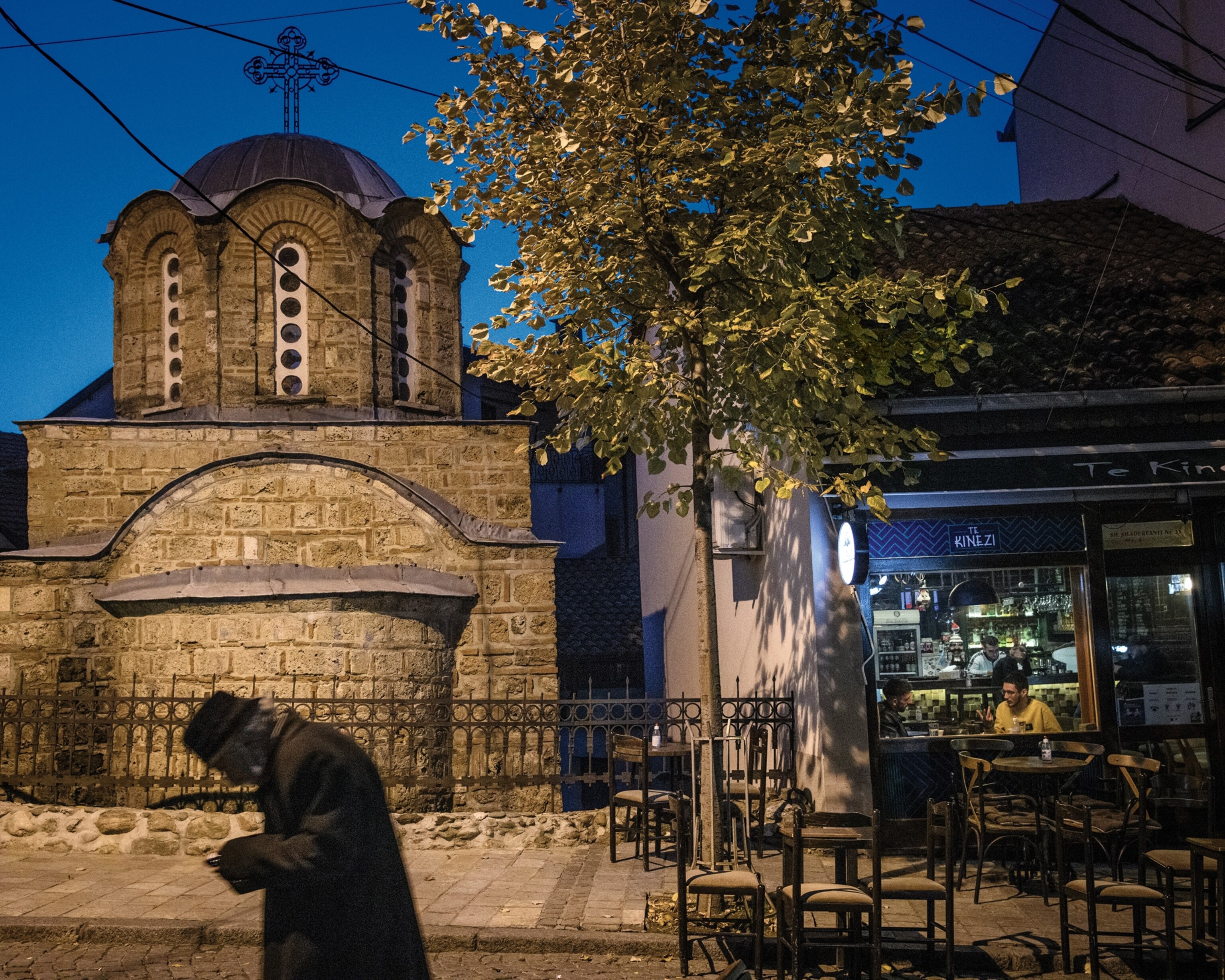 Picture of a man walking in front of a church that is next to a small store, in the late evening.