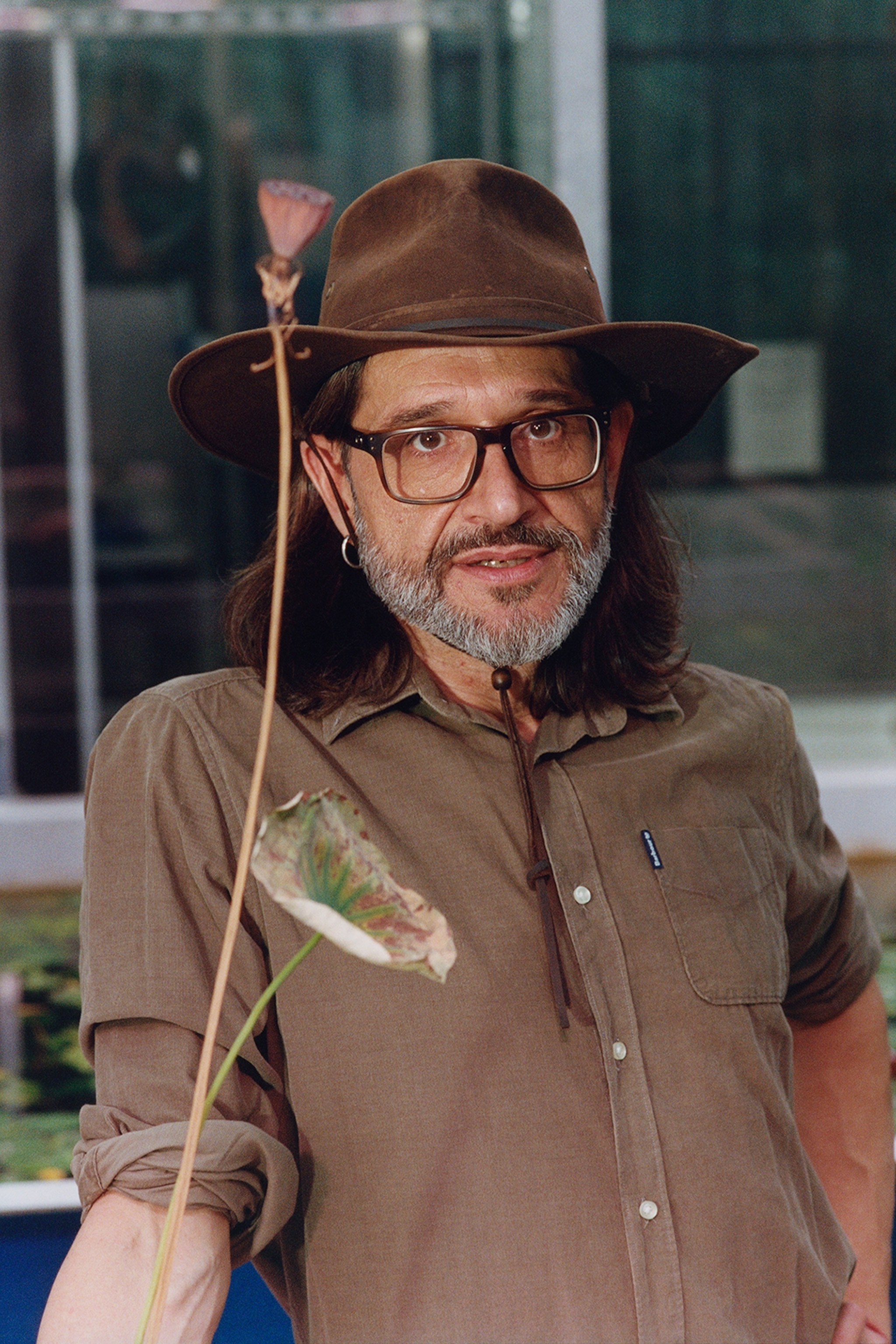 A man stands for a portrait. He is wearing a brown button down shirt and a brown hat. There is a flower in front of him.