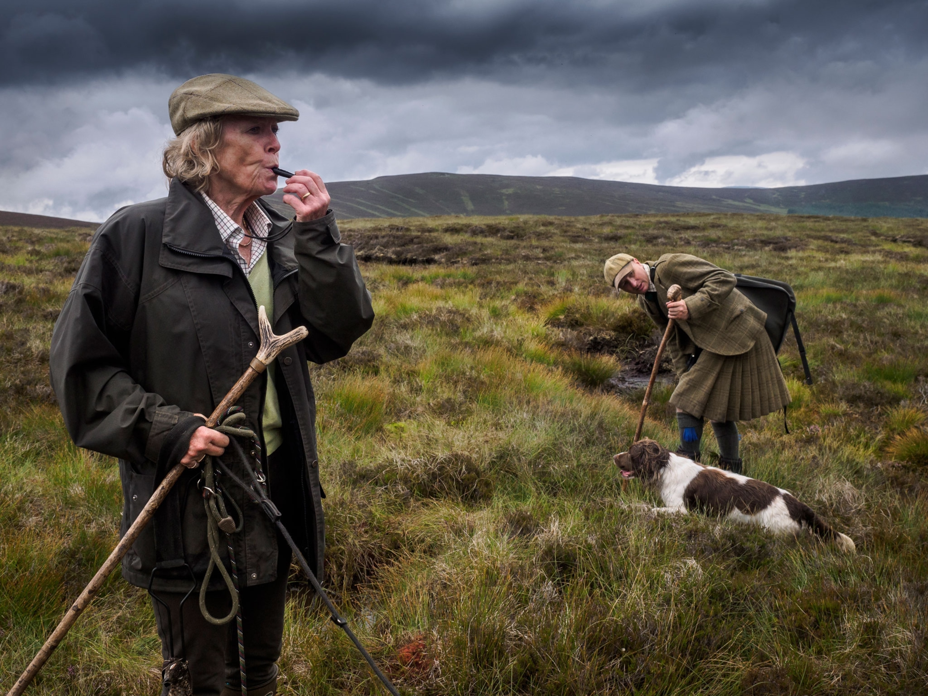 A woman blows a whistle while a man in a kilt looks at a dog during a hunt.