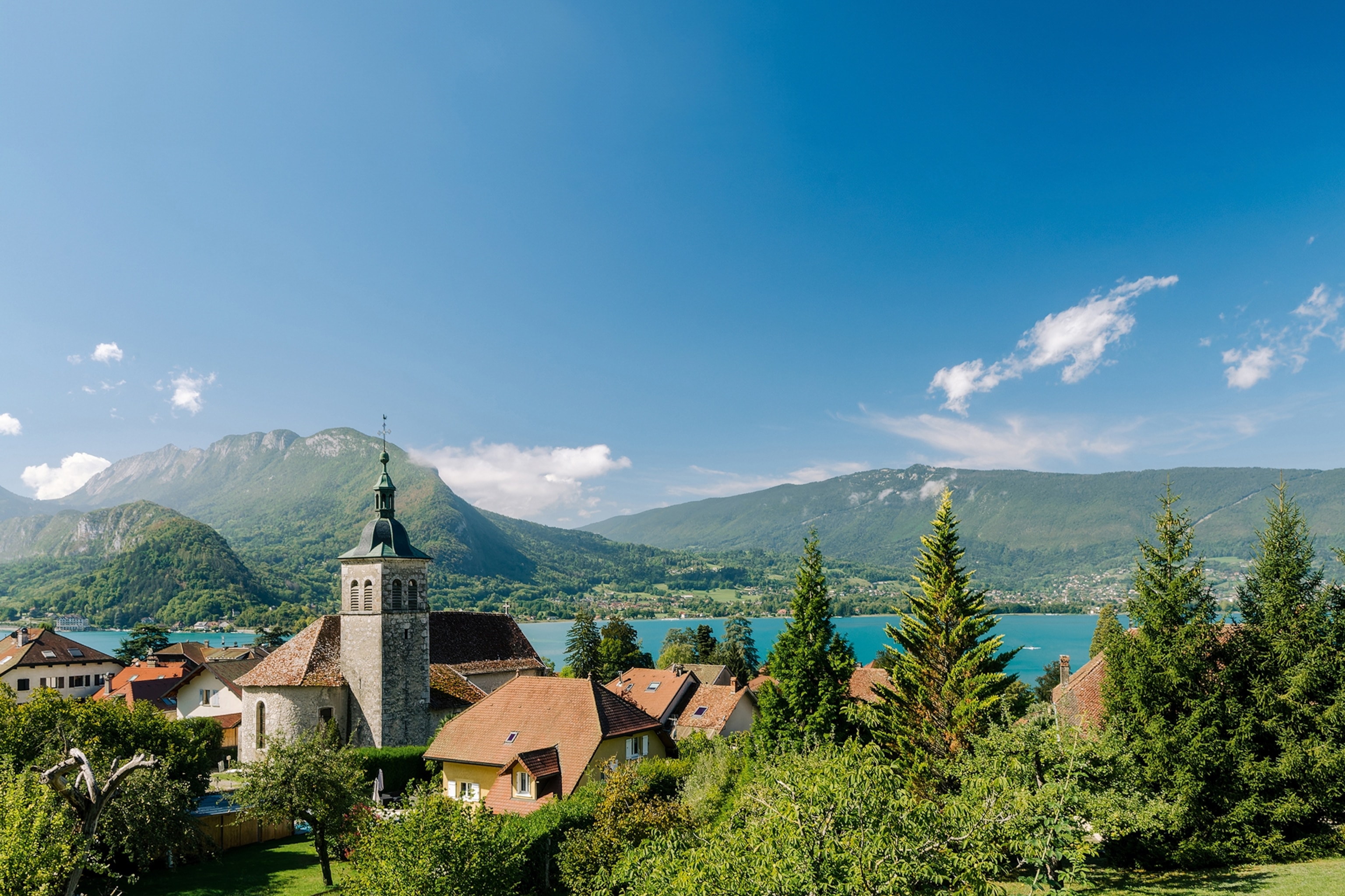 A shot of old French houses with red brick roofs and a lake and mountains in the background.