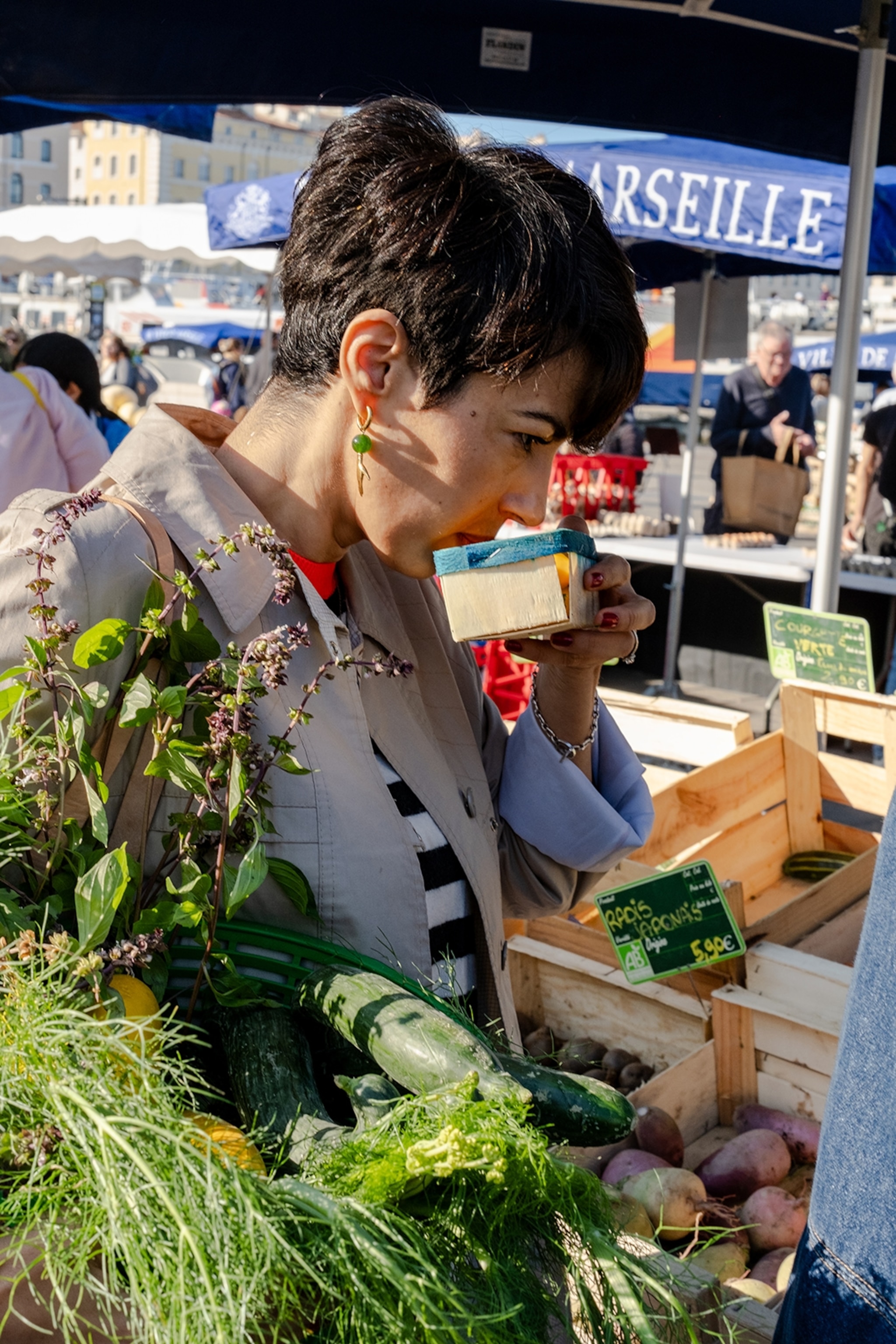 A short-haired woman sniffing at a spice box on a market, holding a bouquet of greenery and vegetables.