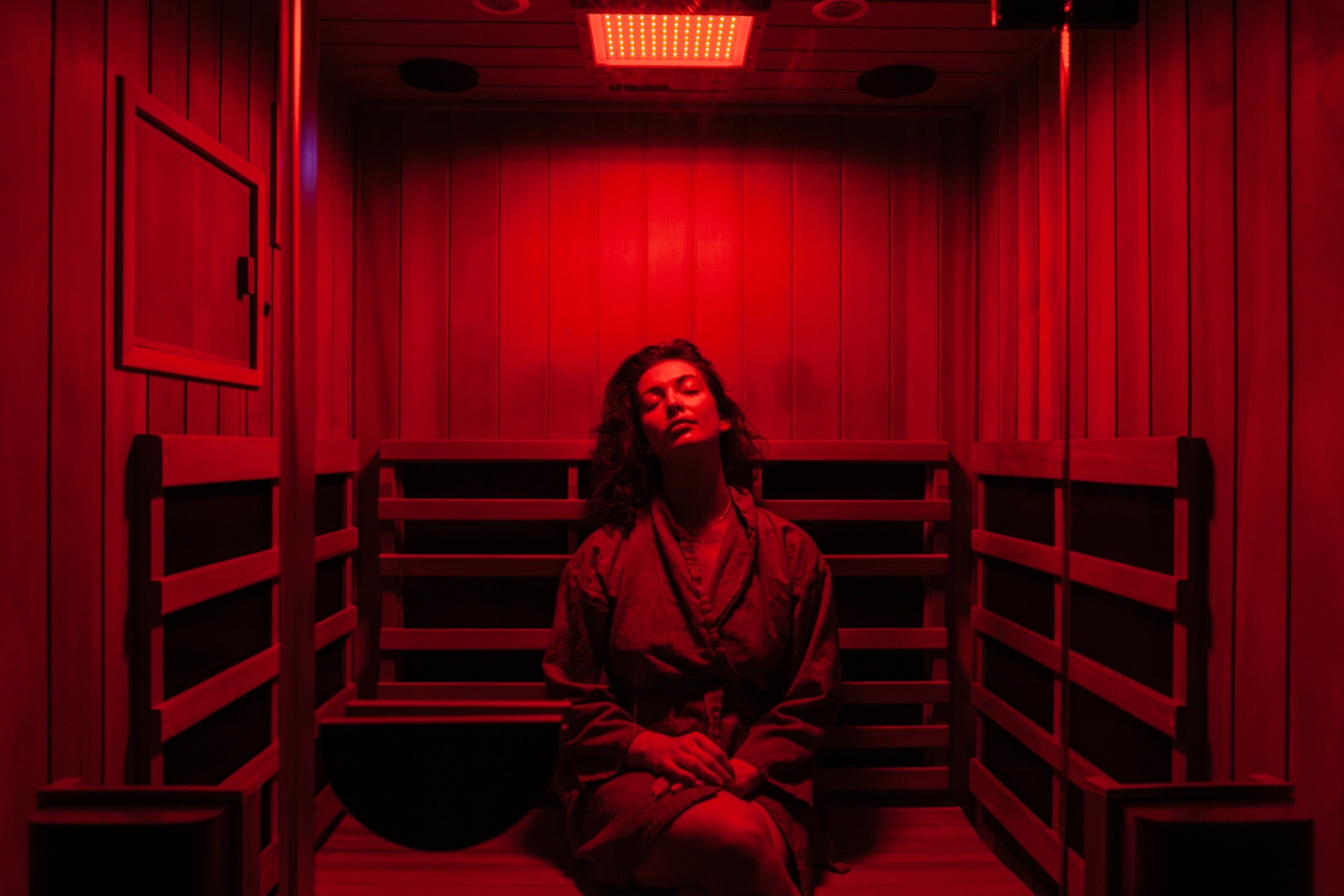 A woman in a robe sitting with eyes closed inside a wooden sauna room that is lit by red light.