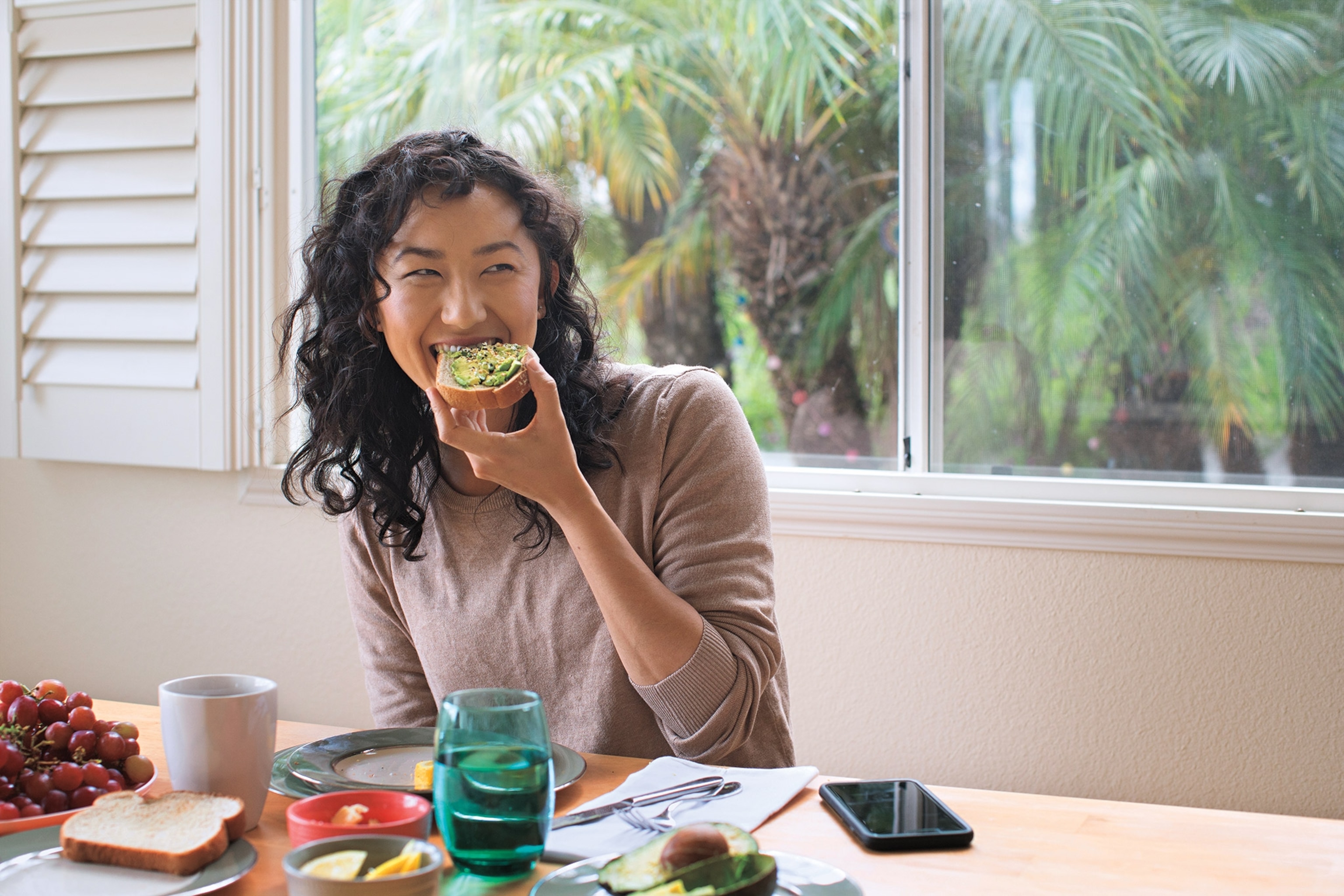 Woman has toast with avocado on at a table with grapes and a phone in front of her.