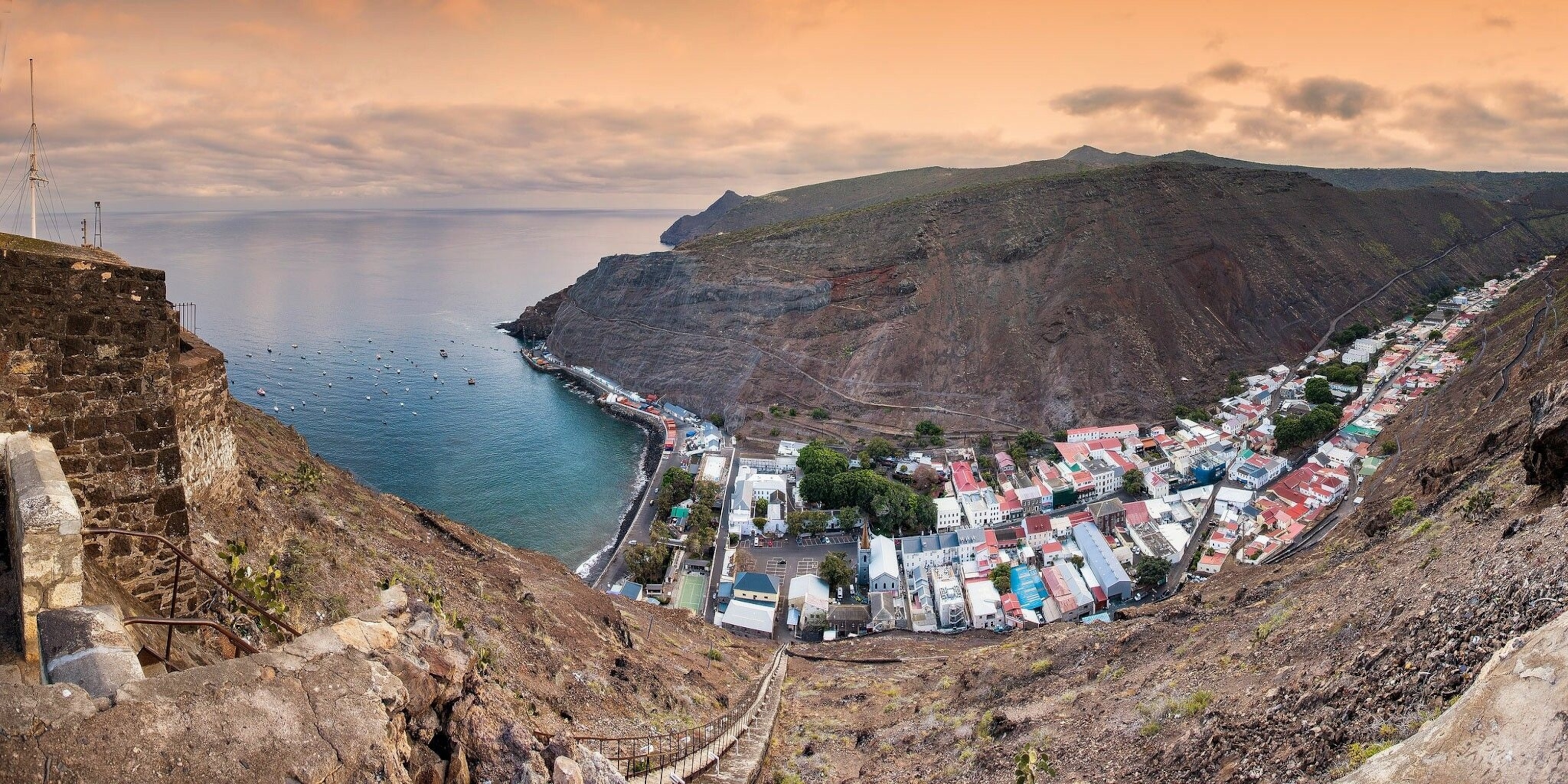 An arial view of Jamestown. Its main road is parallel to the ocean. The town is perpendicular to it, and stretches down a narrow valley between two cliffs, which are brown.