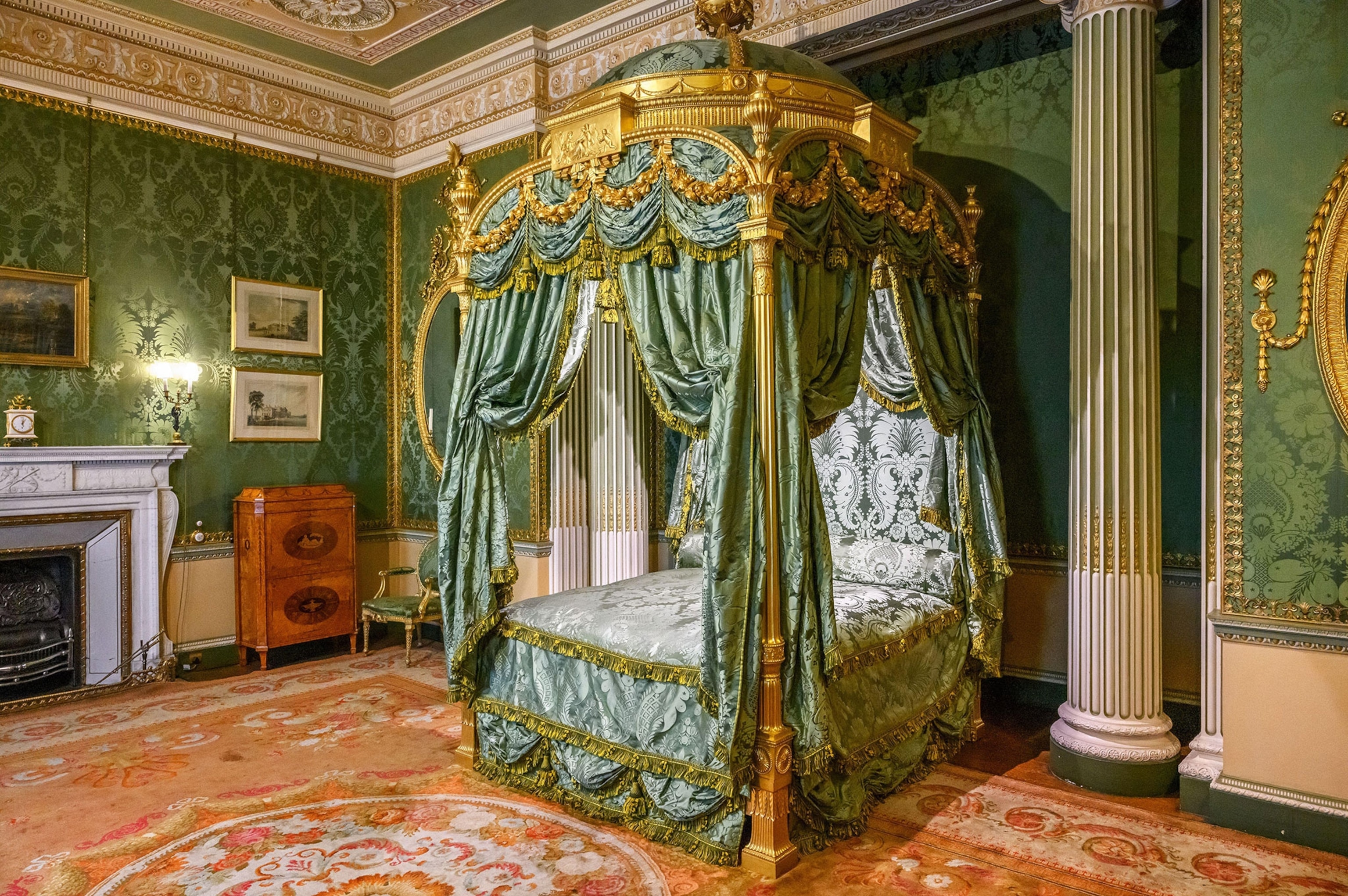 An elegant, old english style bedroom with a four post bed, fireplace, elaborate green wallpaper white marble-like columns