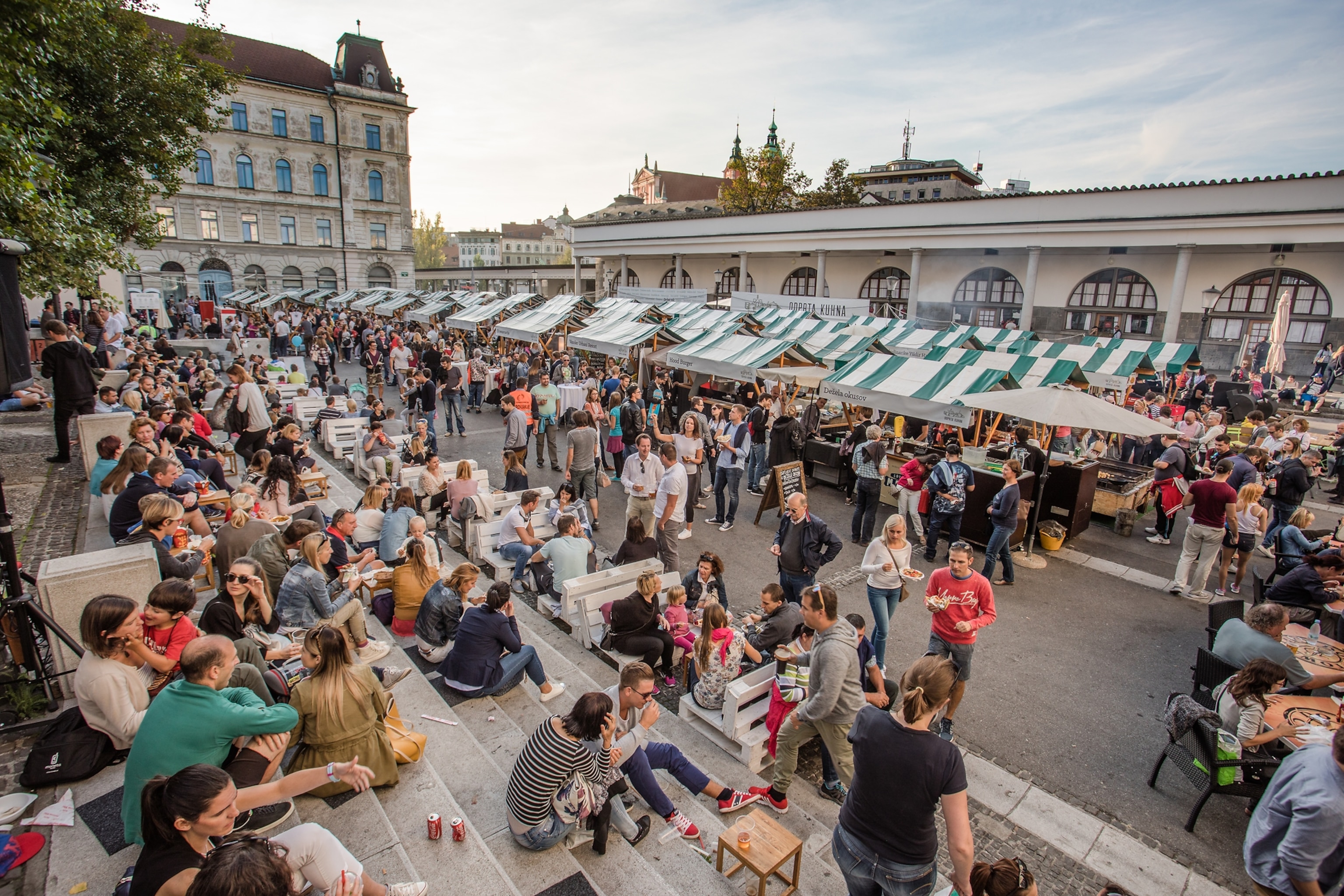 People fill tables and sit on steps in an outdoor market near food stalls selling produce and cooking food.