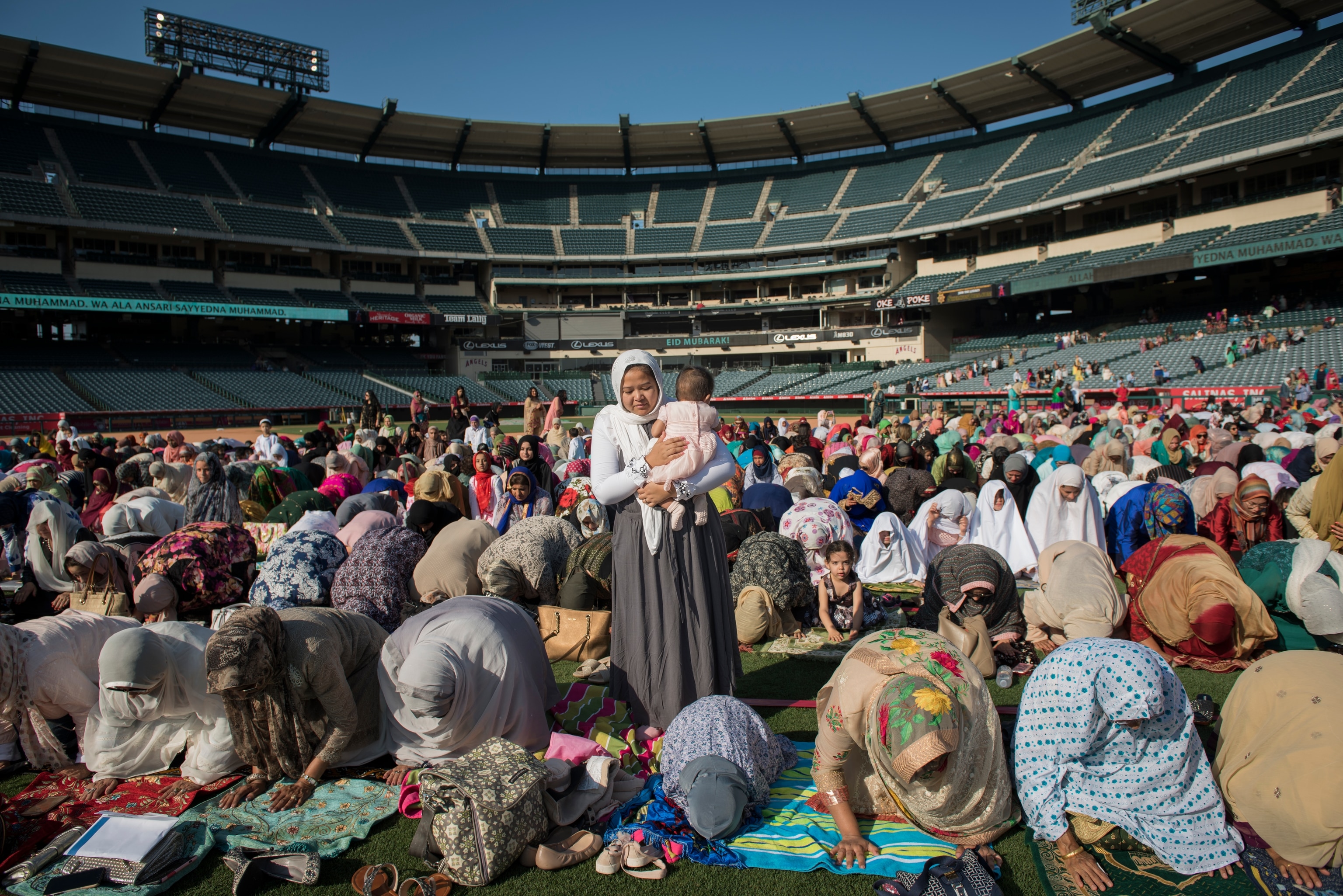 A young Asian woman, wearing a head scarf and holding a baby, stands amid a large group of women and children kneeling, bent over in prayer, in the middle of a grassy field in a large sports arena with empty seats.