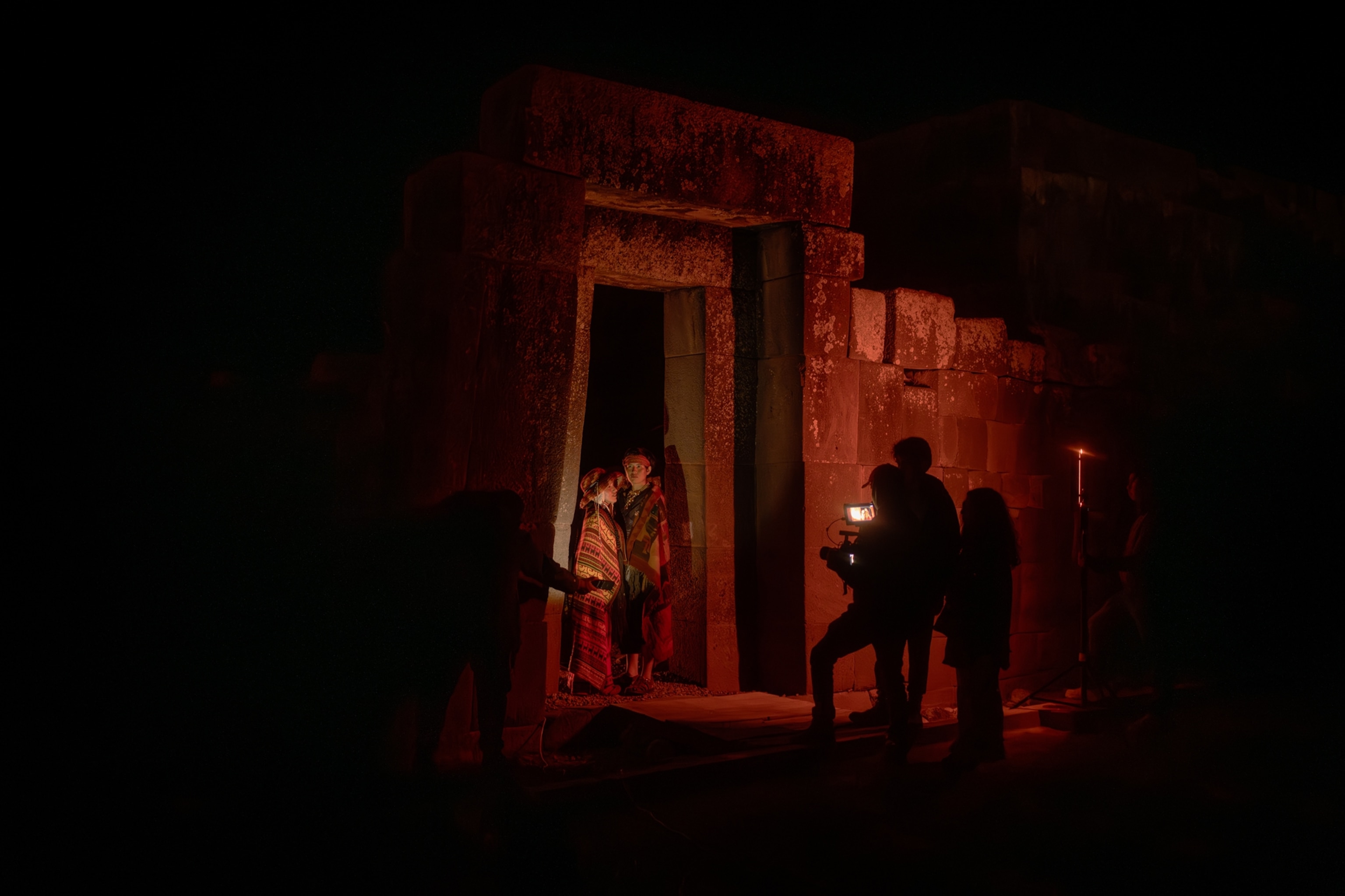 Two figures are seen standing in an open stone doorway, at the Ushnu. The lighting is red. Silhouettes are seen on the right side of the image with a videocamera.