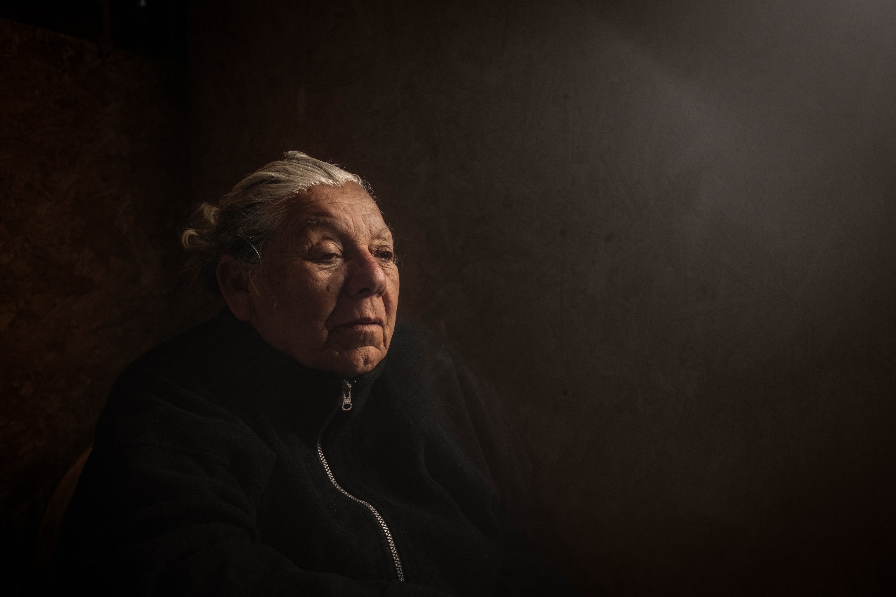 a side portrait of an older woman. Lighting is dark, and her grey hair is pulled back.