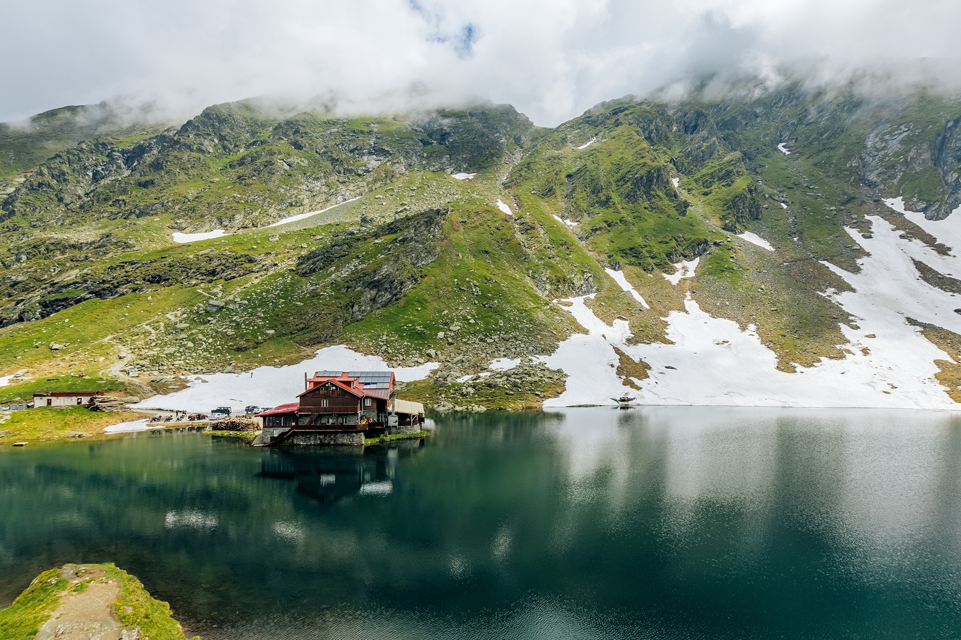 A view across a mountain lake with a hut in the left hand side and an out-of-sight mountain in the background.