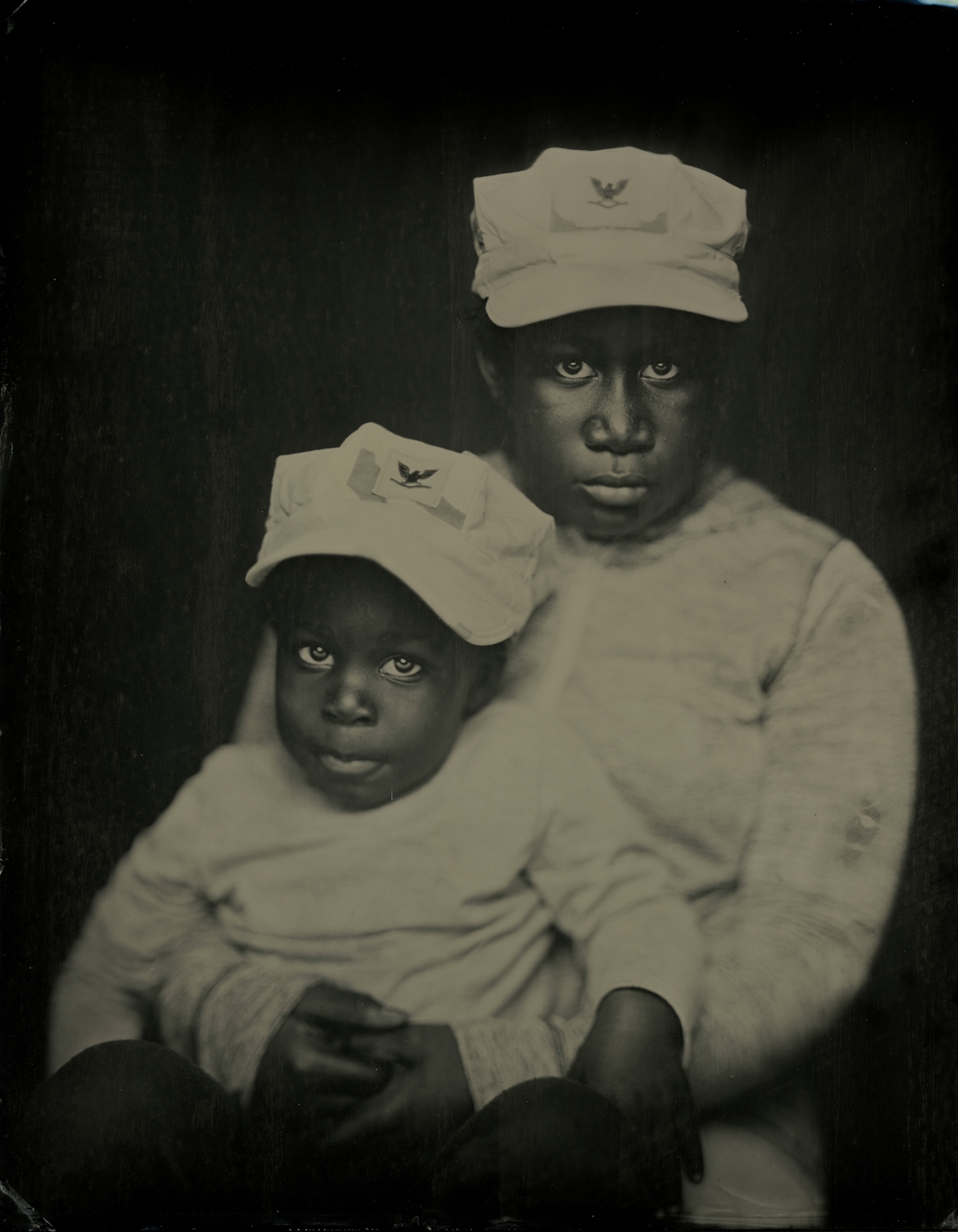 Young boy holding baby on lap posing portrait, both wearing their father's military hats