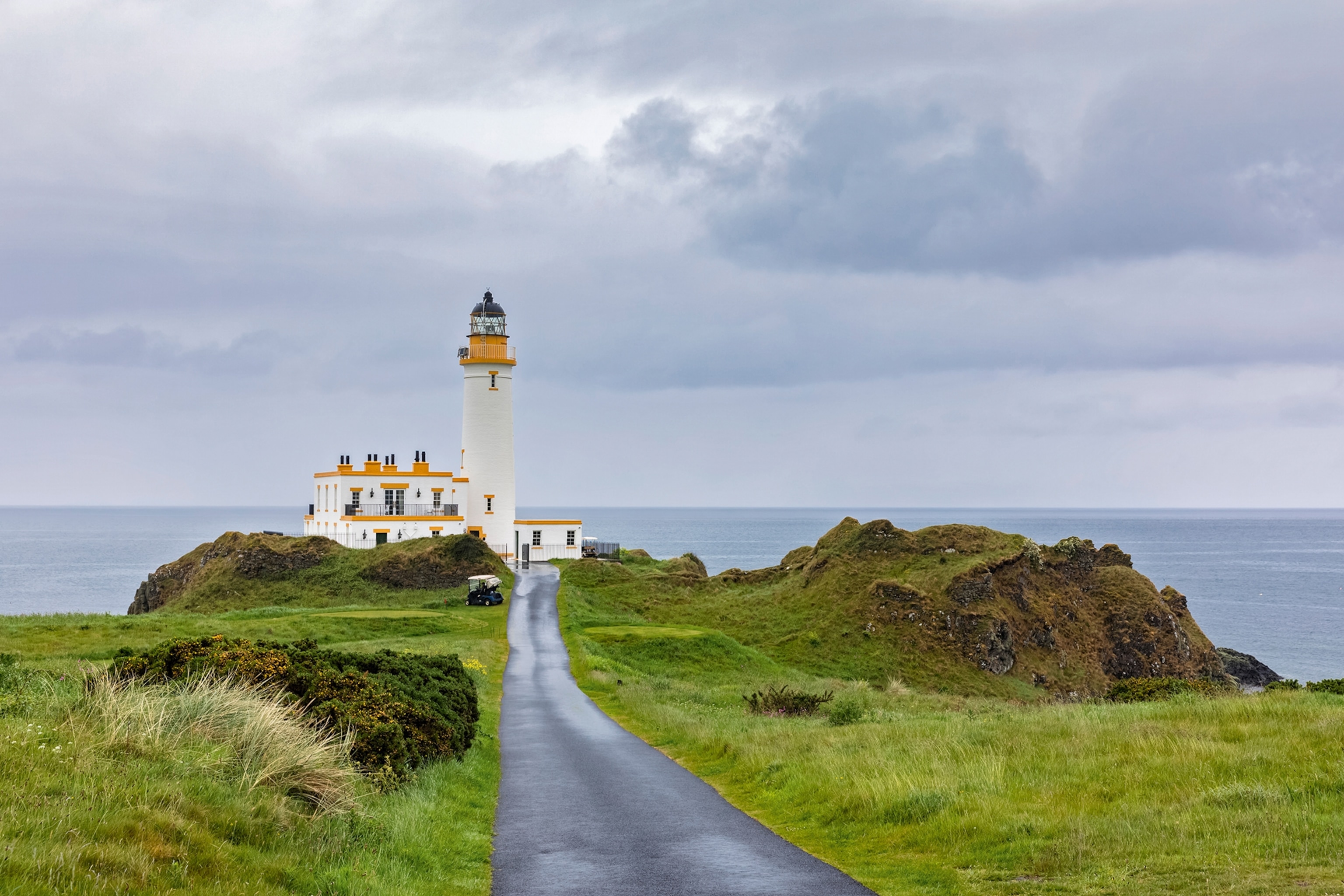 A long road leading down to a white lighthouse on the coast