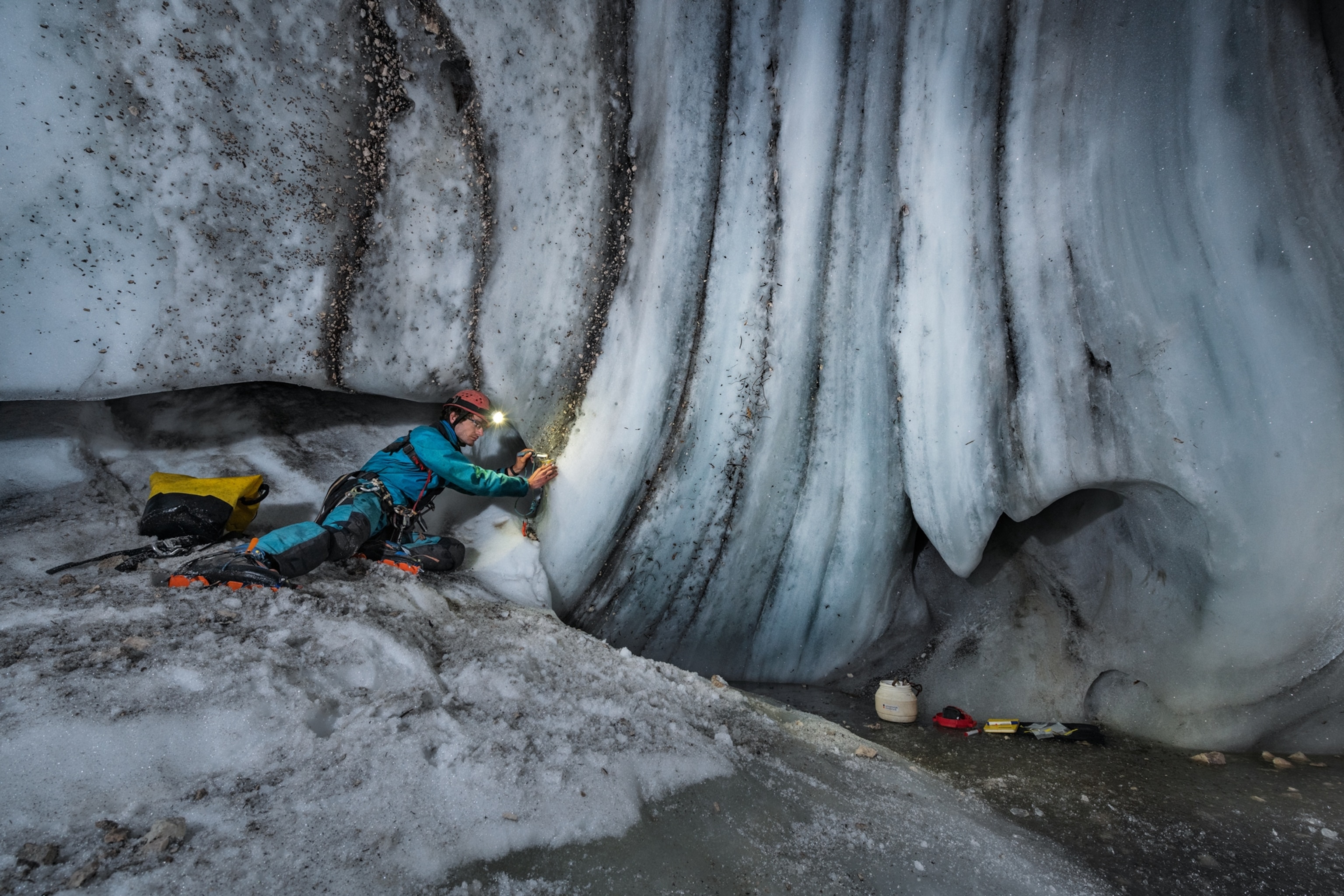Picture of person in a blue suite and red hard hat collecting debris from ice on floor and walls of the cave.
