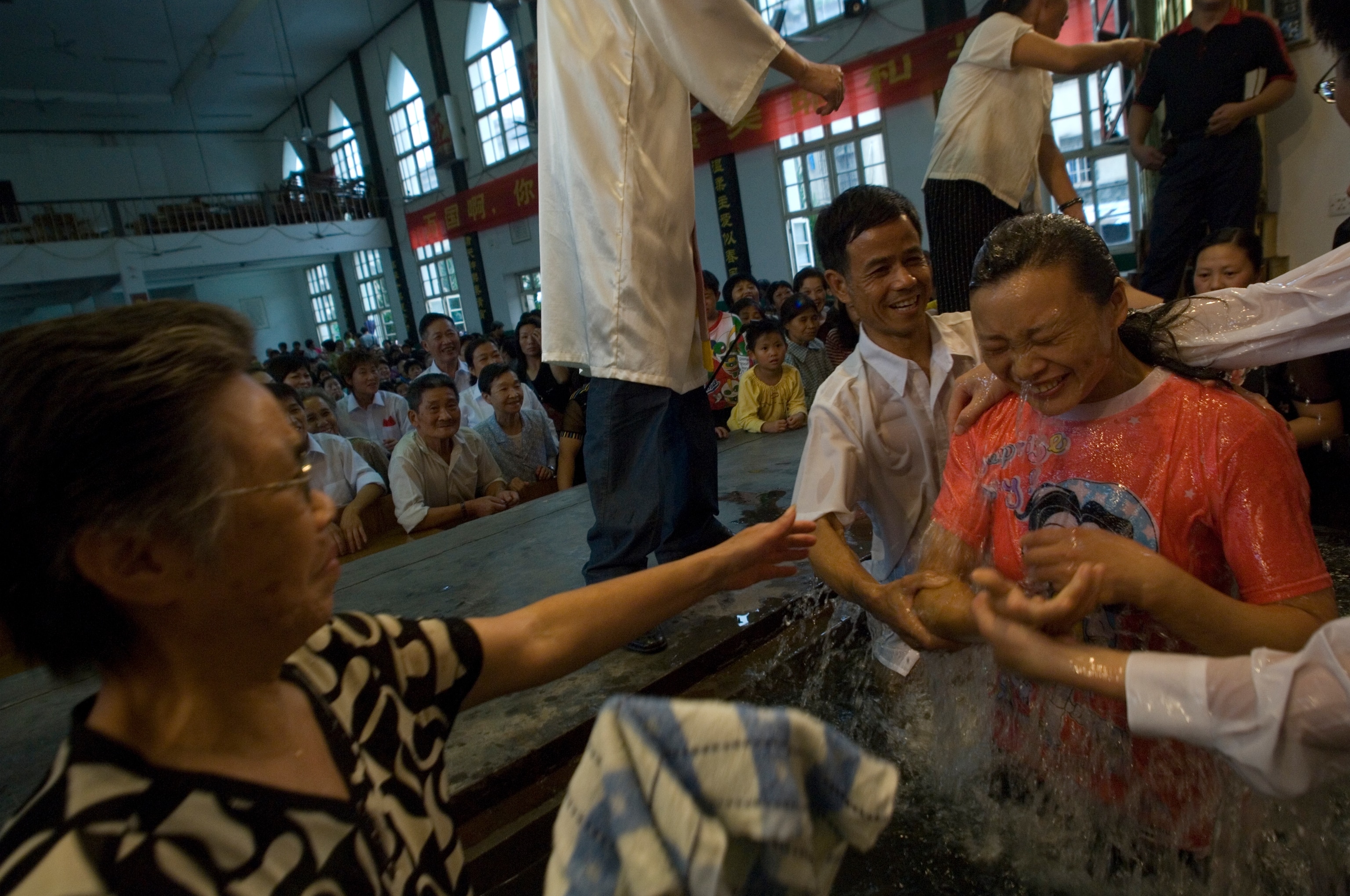 A young Asian woman, smiling with eyes closed and water pouring off her, is pulled out of a tub of water by a smiling Asian man during a baptism ceremony inside a church. A crowd of Asian people sit in pews in the background.