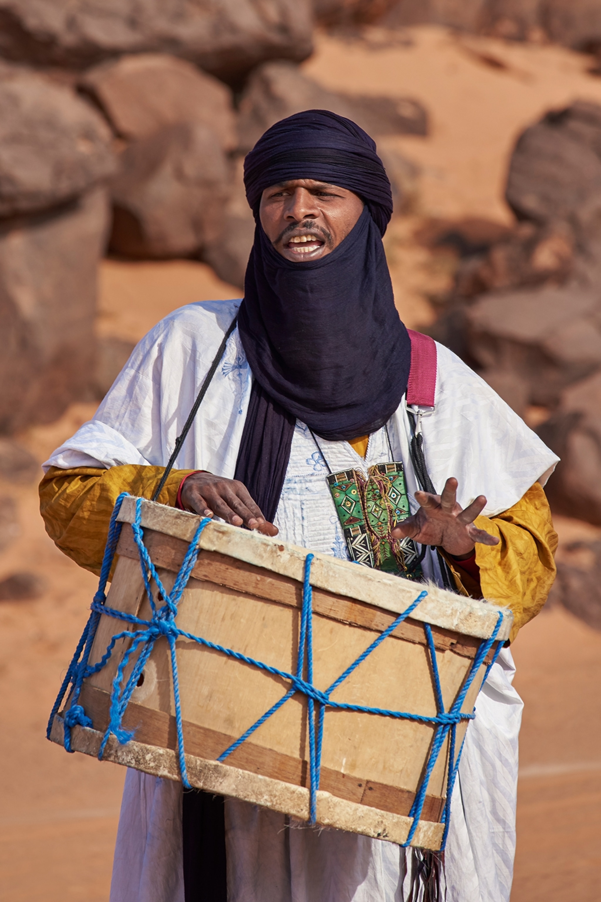 A Tuareg wearing a tagelmoust, a blue headscarf. He is playing a drum which hangs around his neck, attached by a blue rope.
