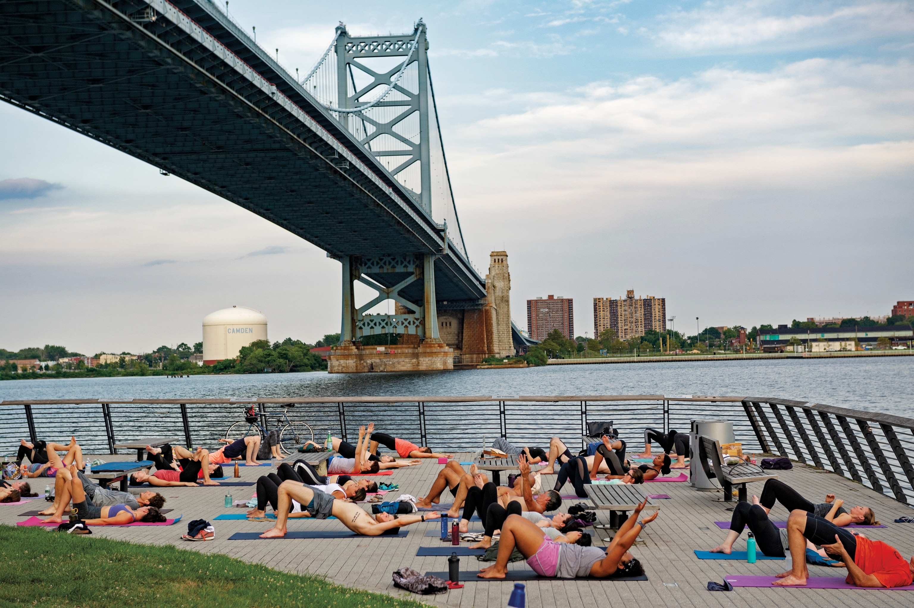 A group of people do yoga in a public city park under a bridge next to a river.