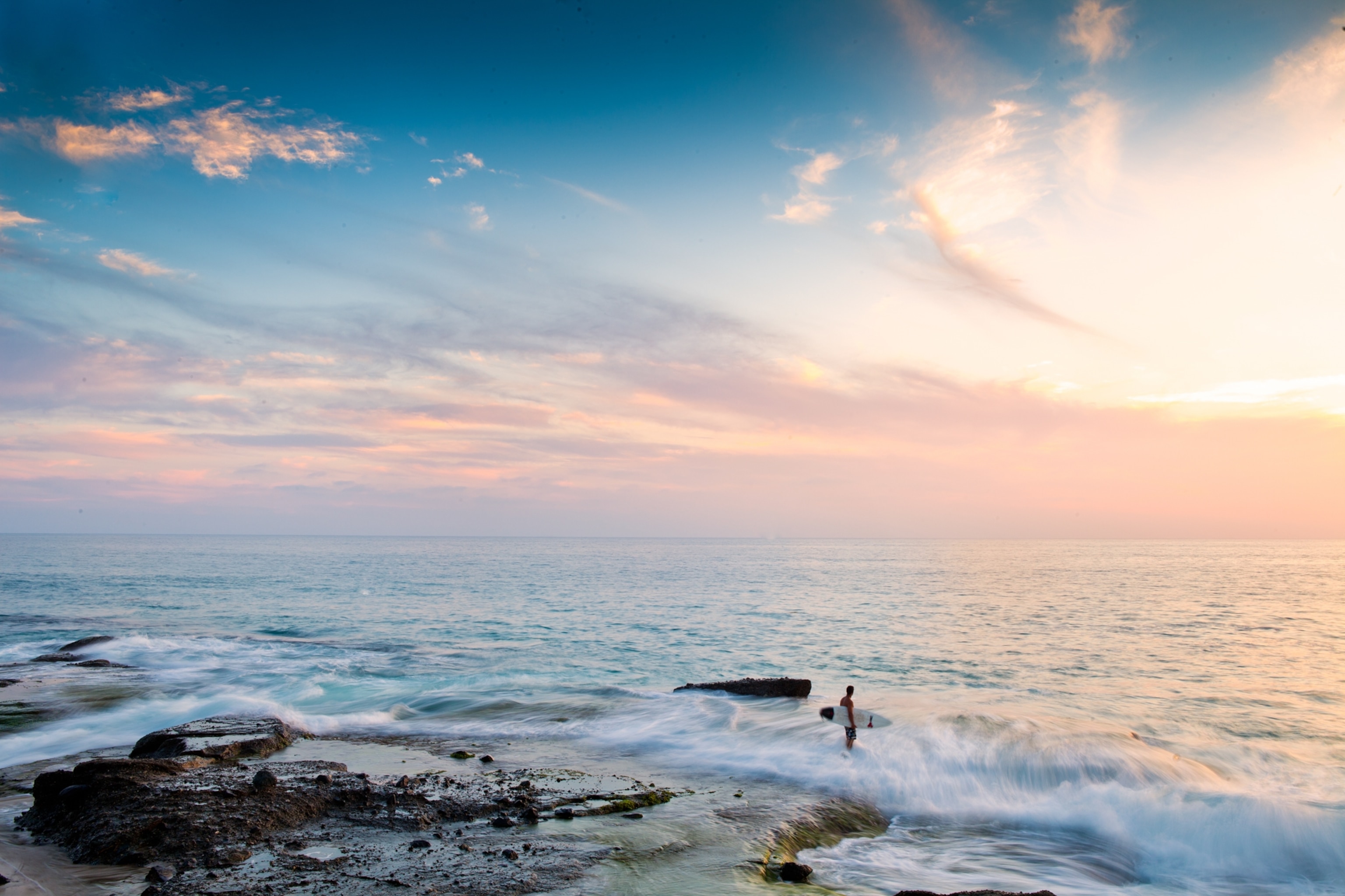 A surfer walking across the reef at sunset to access the waves.