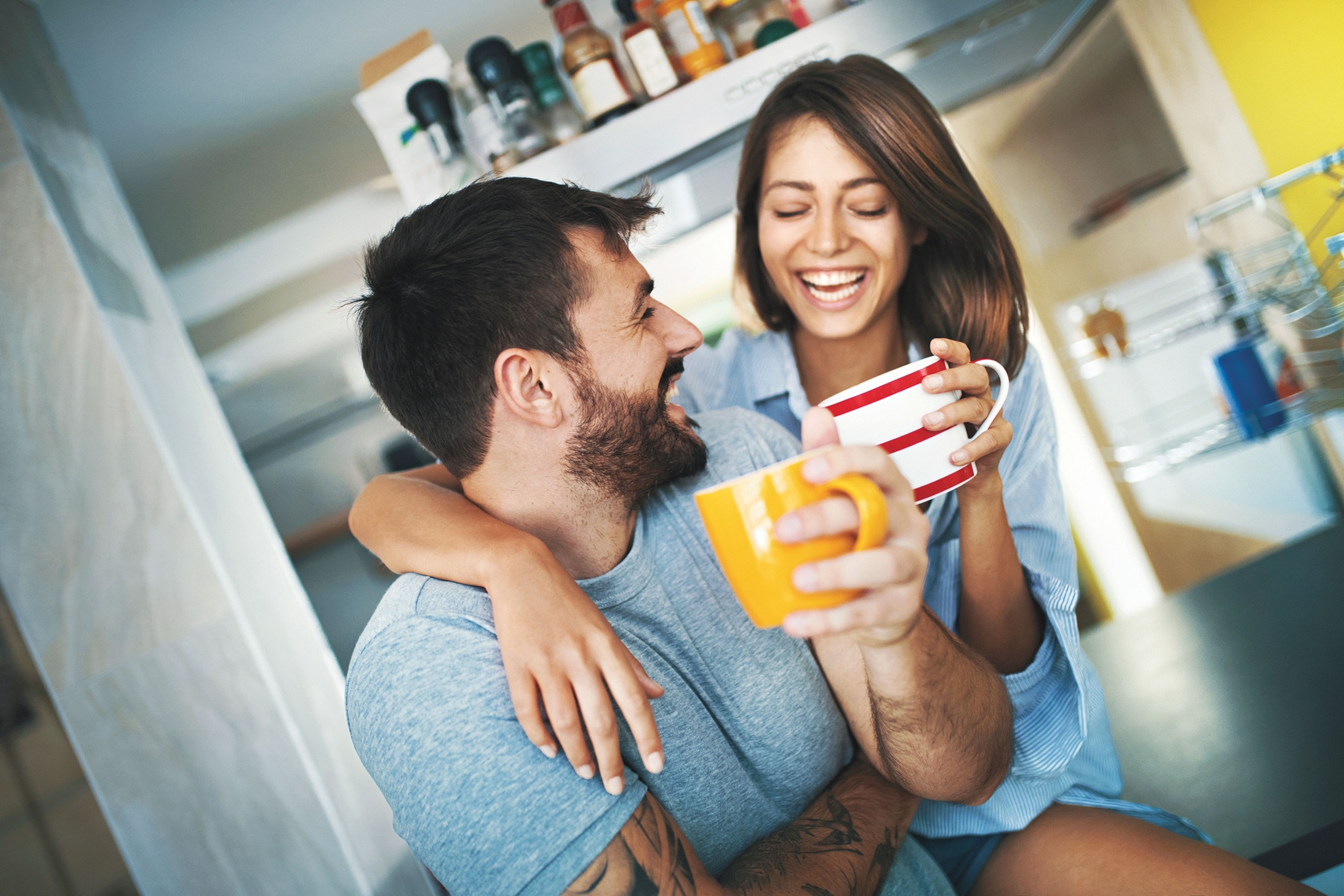 A couple smiles while holding coffee cups in a bright kitchen.