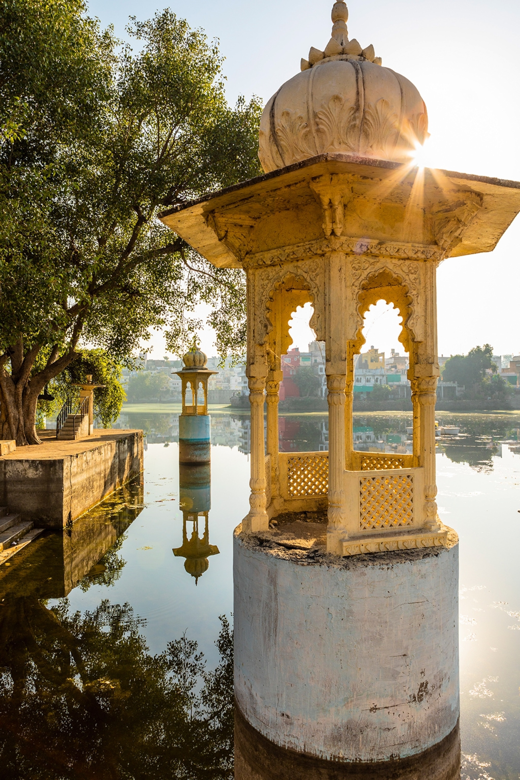 A lake scene in direct sunlight with temple-like structures rising from the water, its mirror on the water surface.