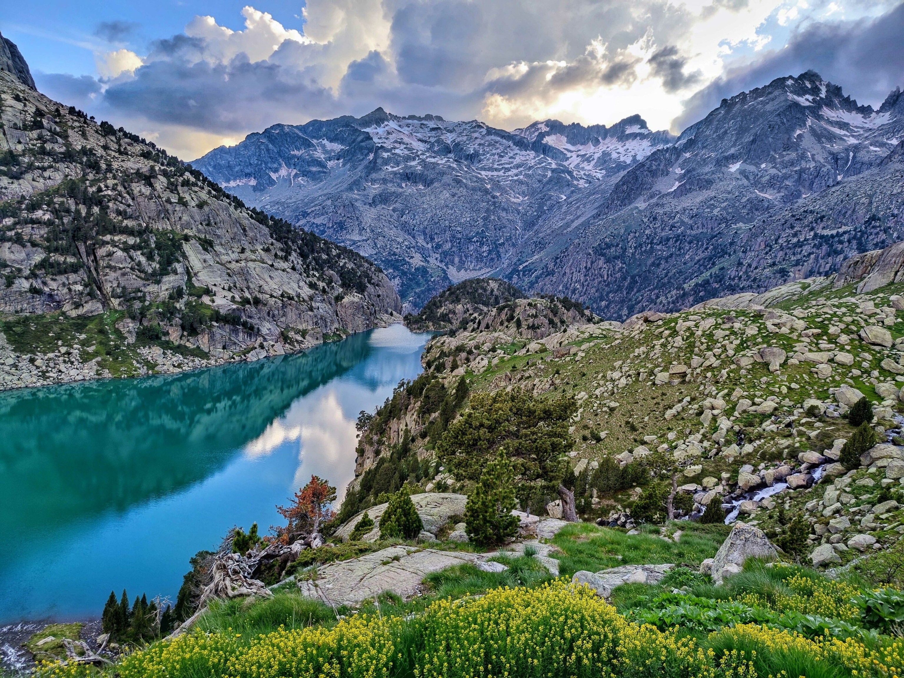 A glassy lake below snow-capped mountains. Greenery surrounds.
