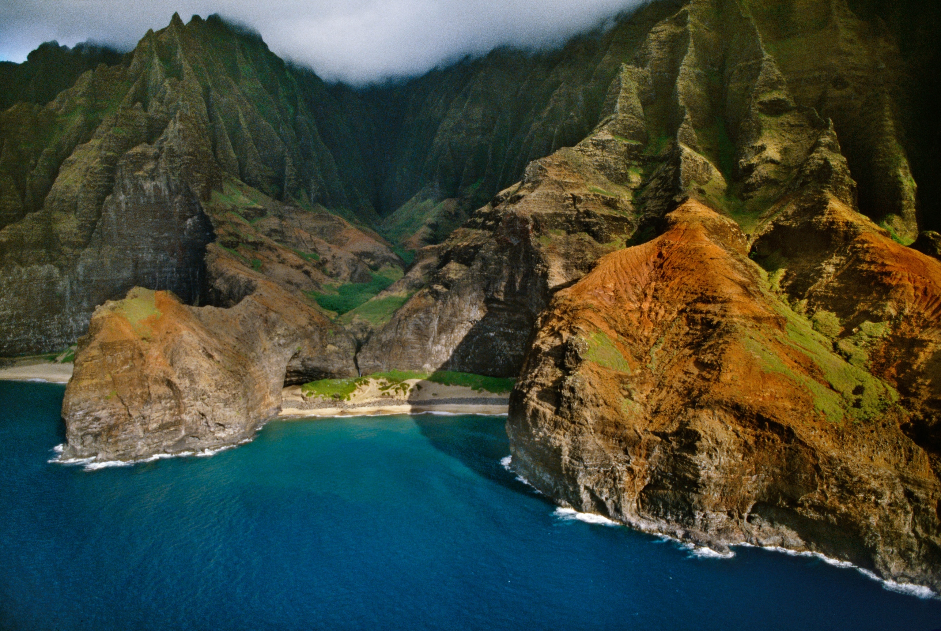 Clouds roll in from atop a green mountains onto the coast.