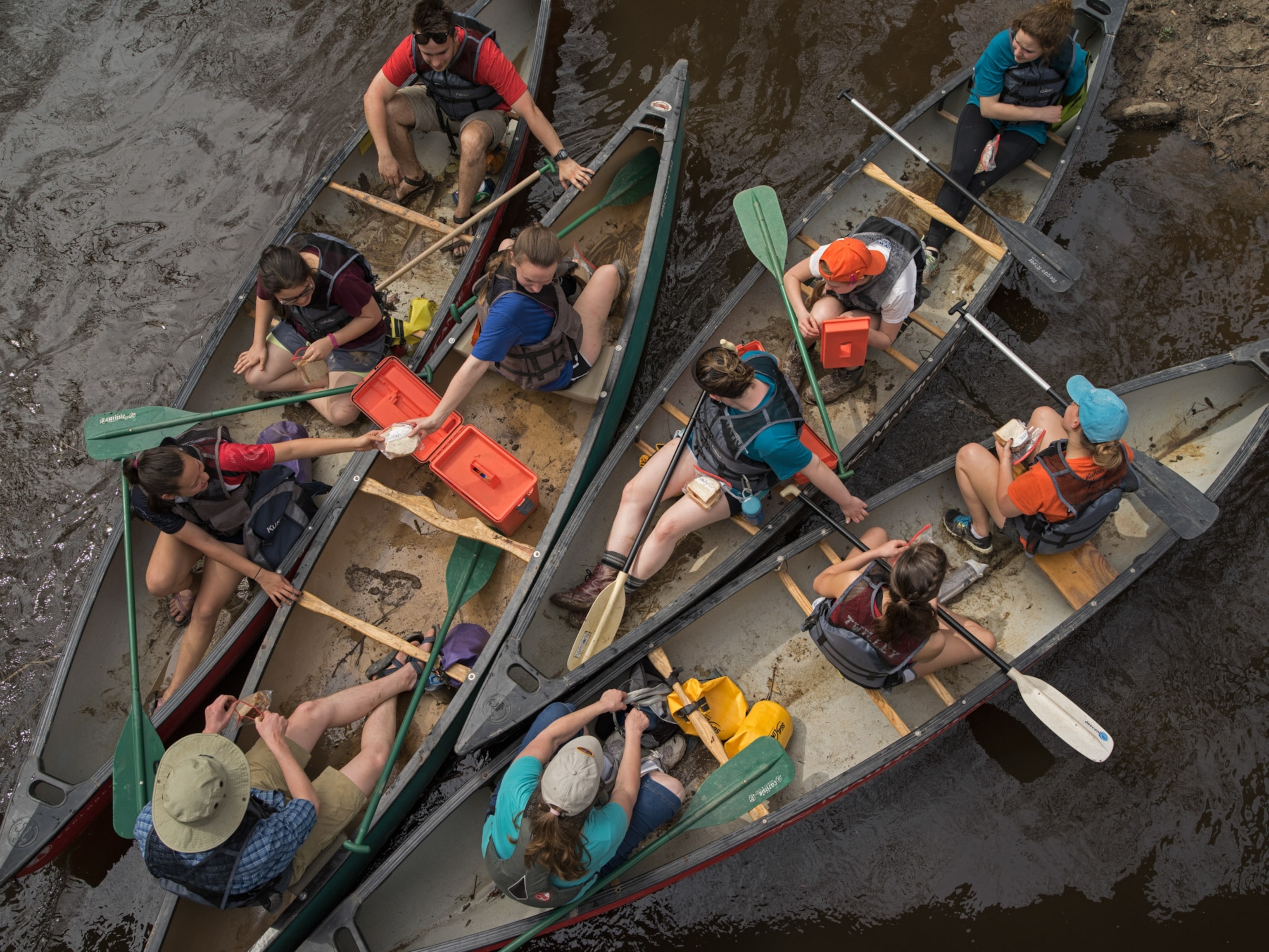 An overhead image of people in canoes some holding paddles and handing lunch to each other.