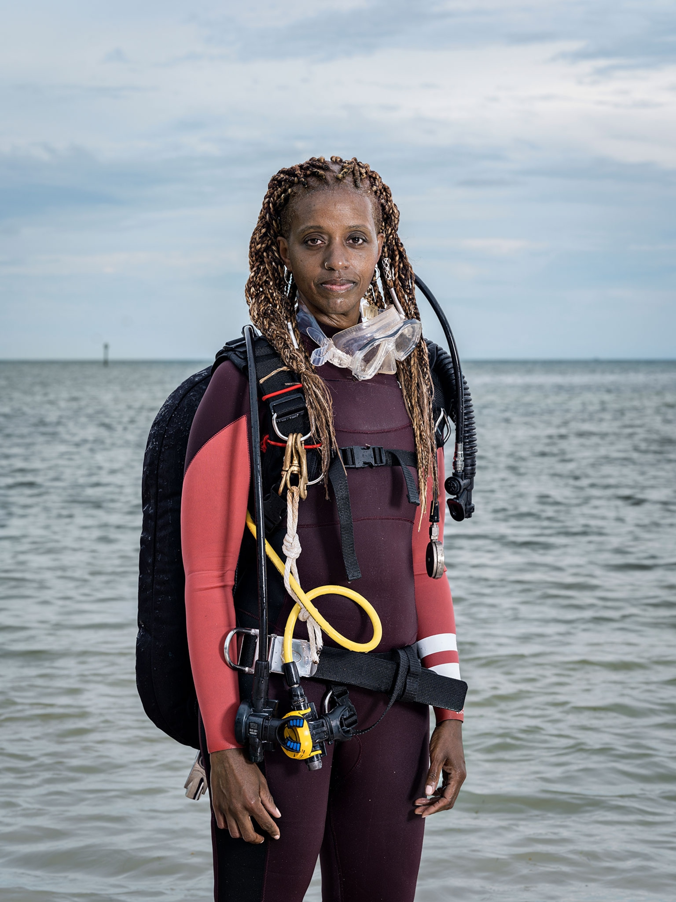 A woman stands waist deep in water wearing scuba gear, goggles hanging around her neck.