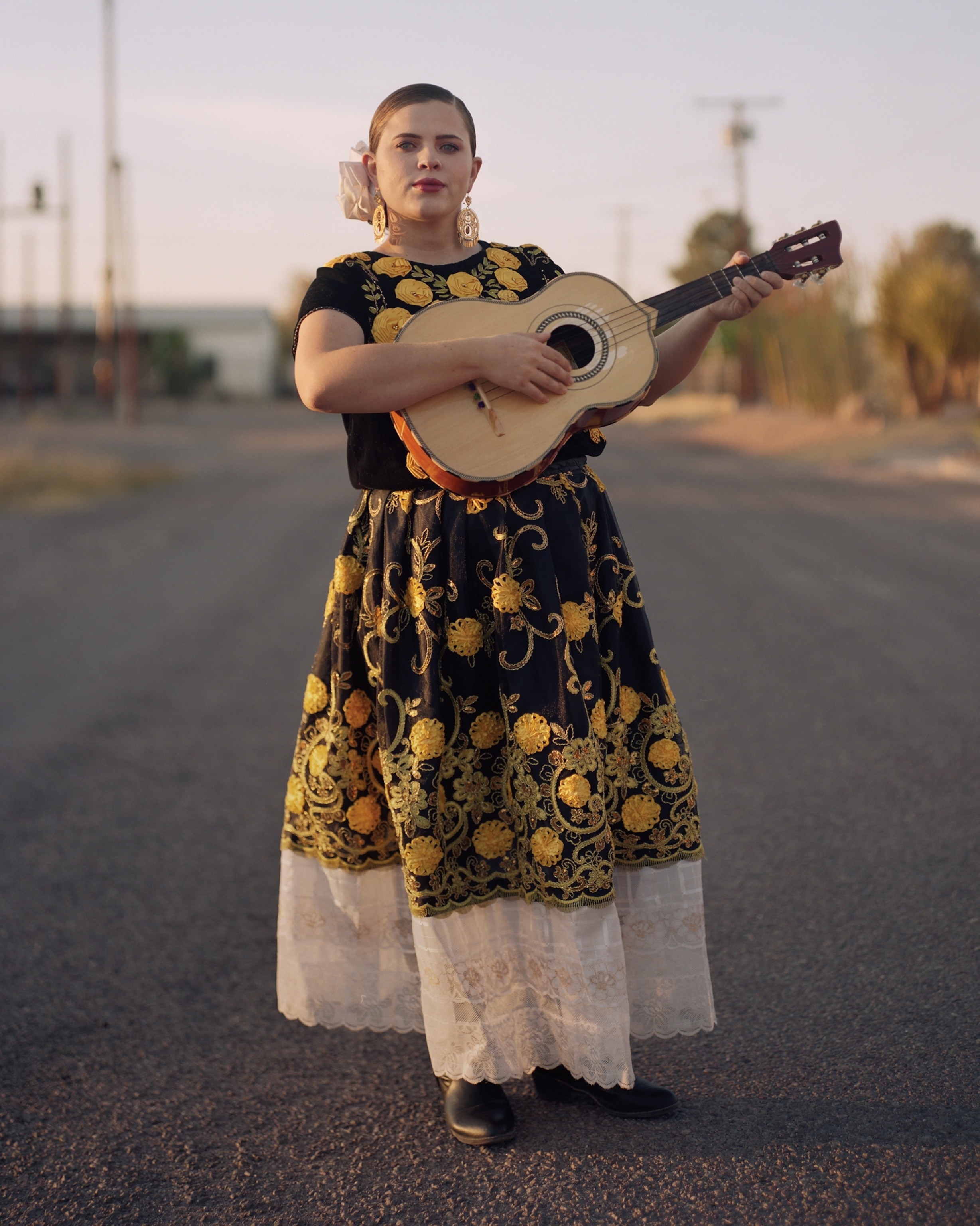 Picture of young woman in long black dress with golden embroidery and white lace standing with guitar.
