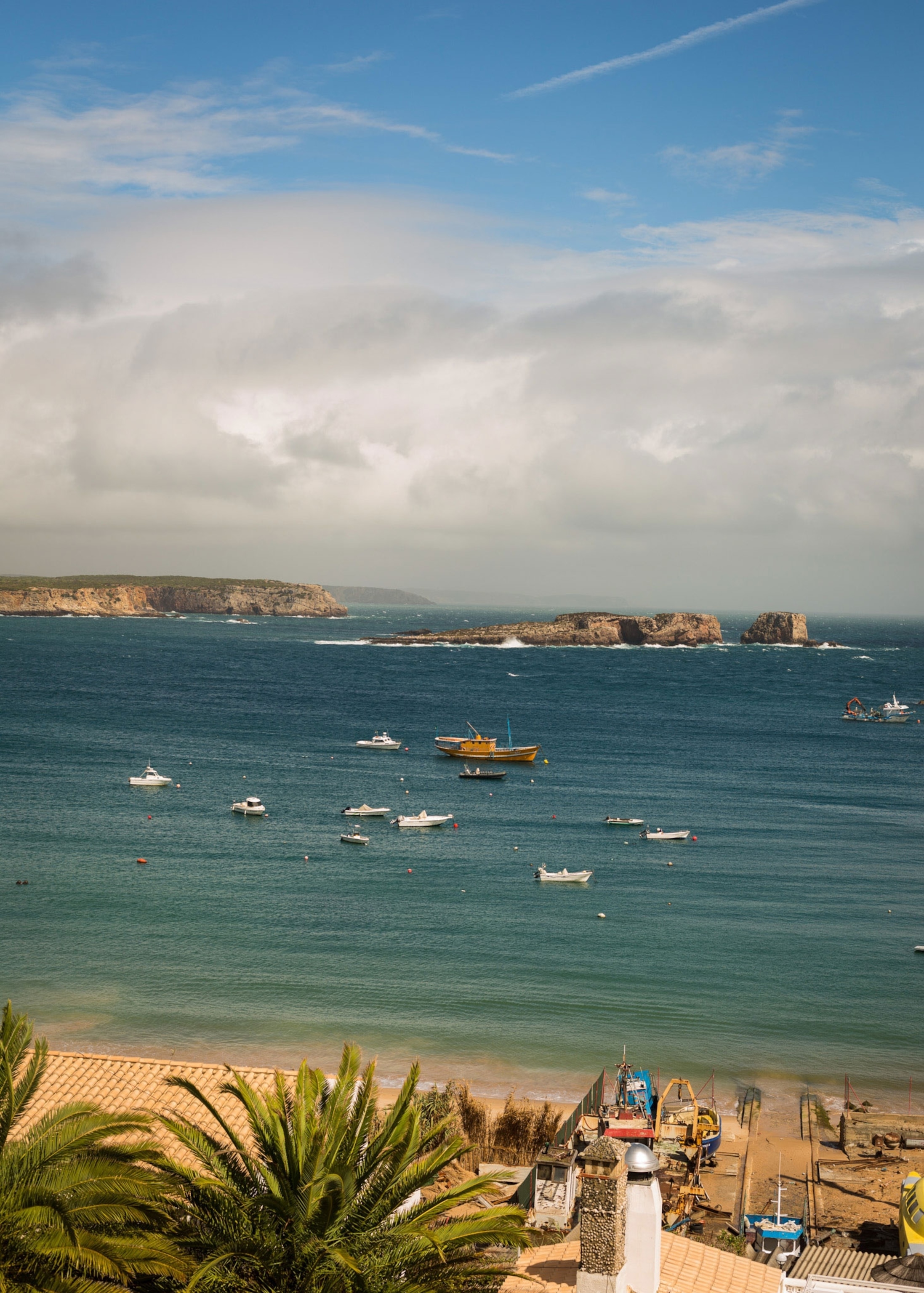 The sand meets the blue ocean with a cluster of boats sitting in front of large rocks in the water.