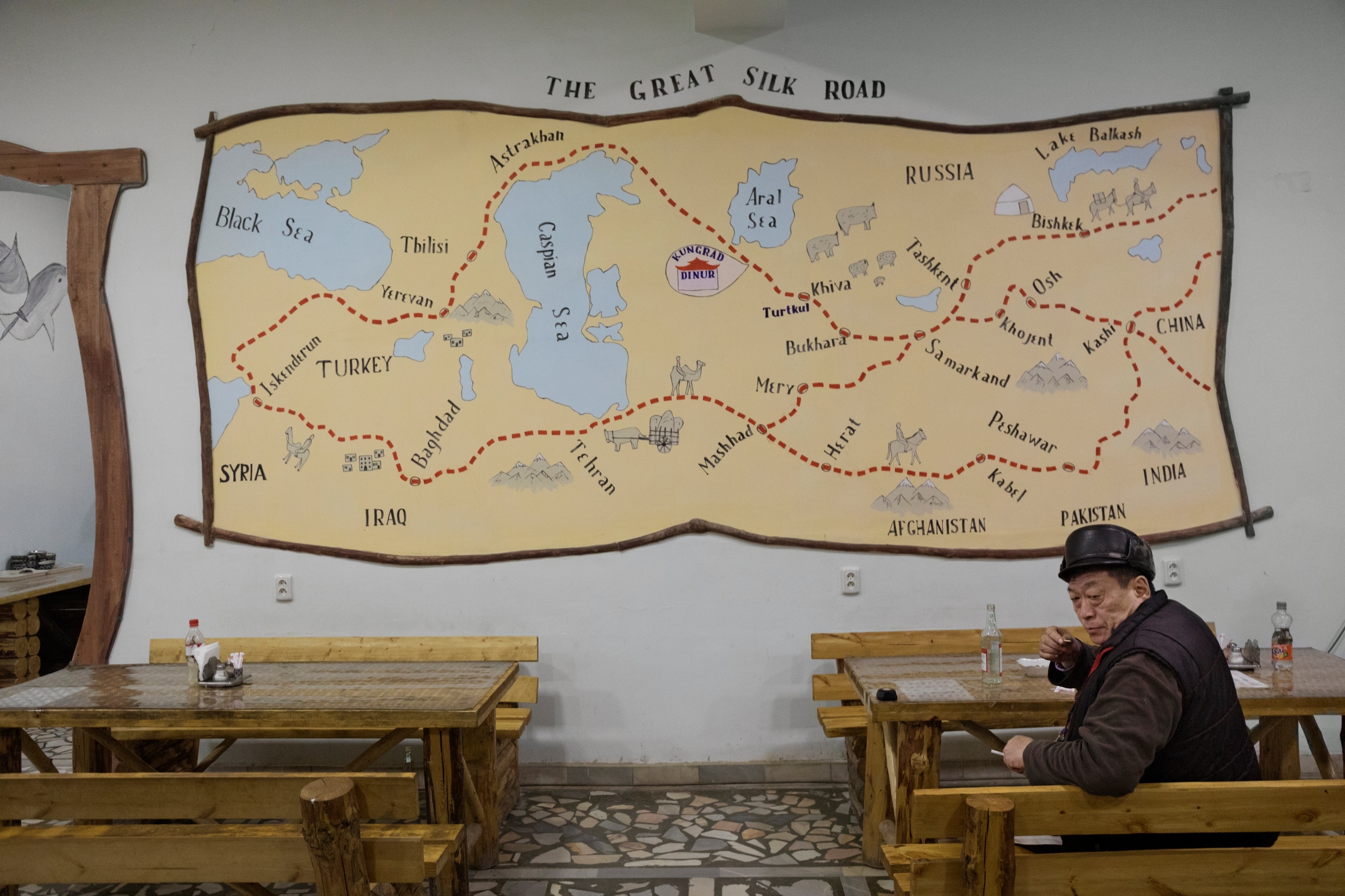 A man sits at a wooden table in a restaurant displaying a large map of the Silk Road on the back wall.