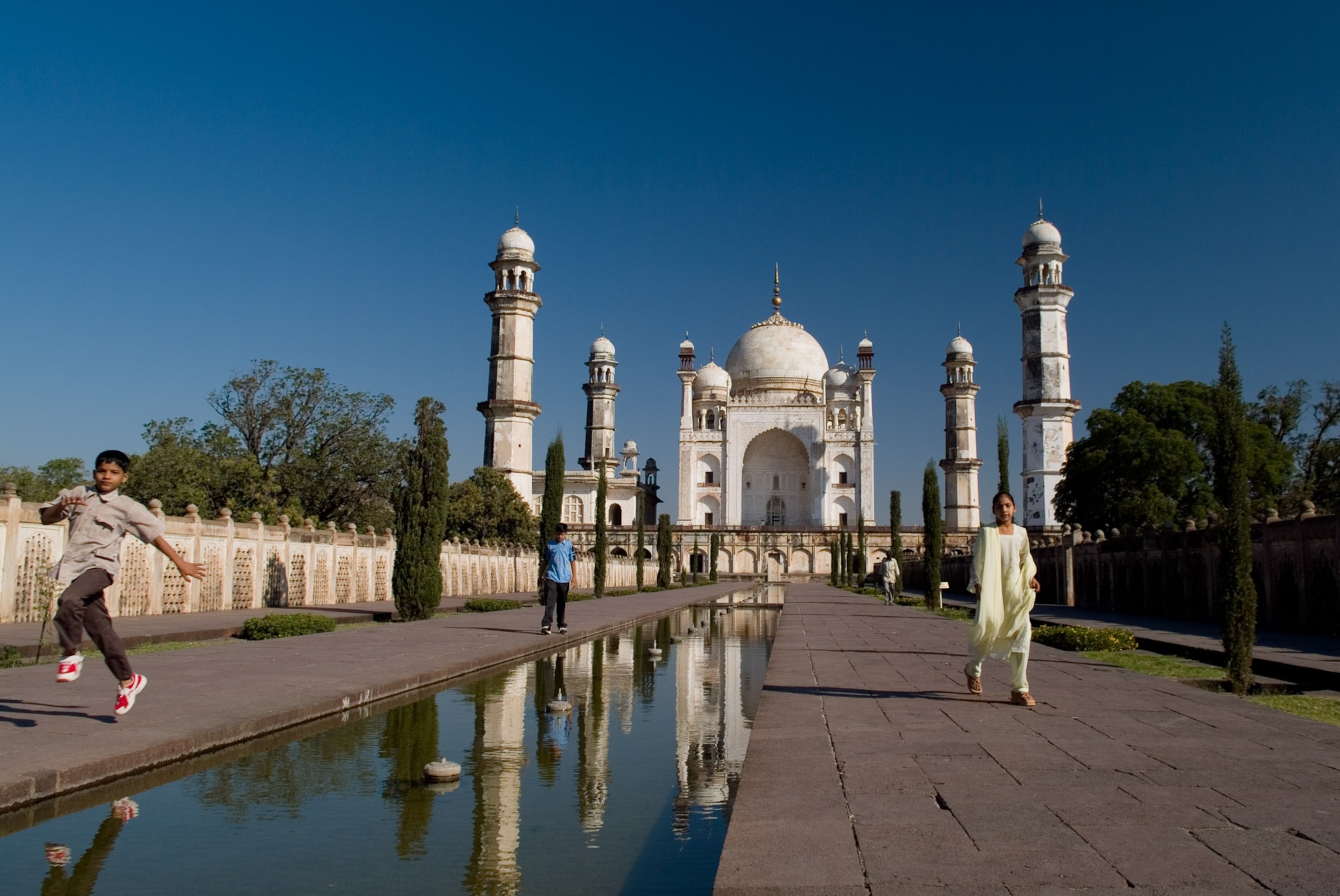 A mosuleum white under a blue sky, a child jumps over a pool of water.