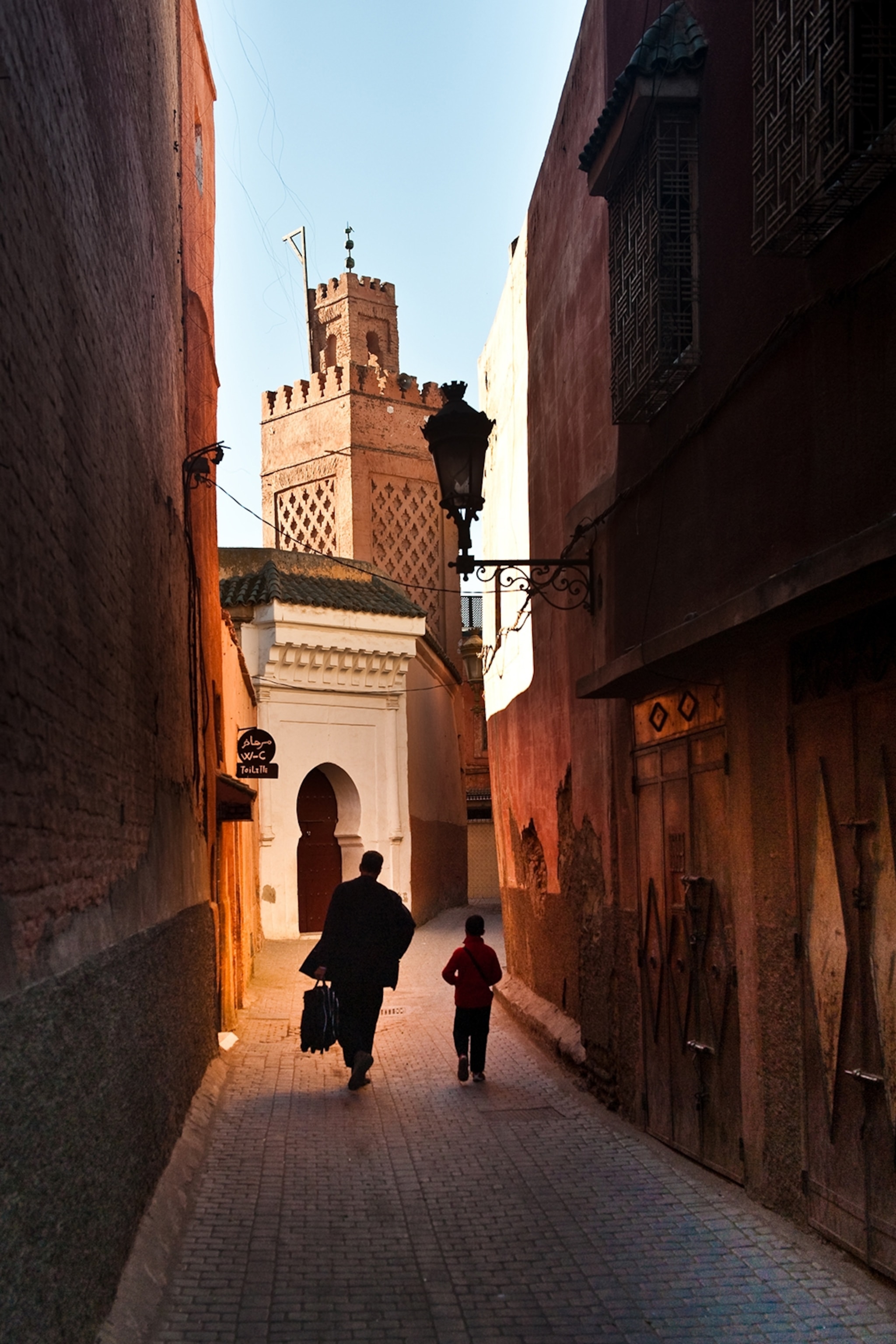A child and adult walk through the dark streets of marrakech. The tall walls don't allow sunlight to enter